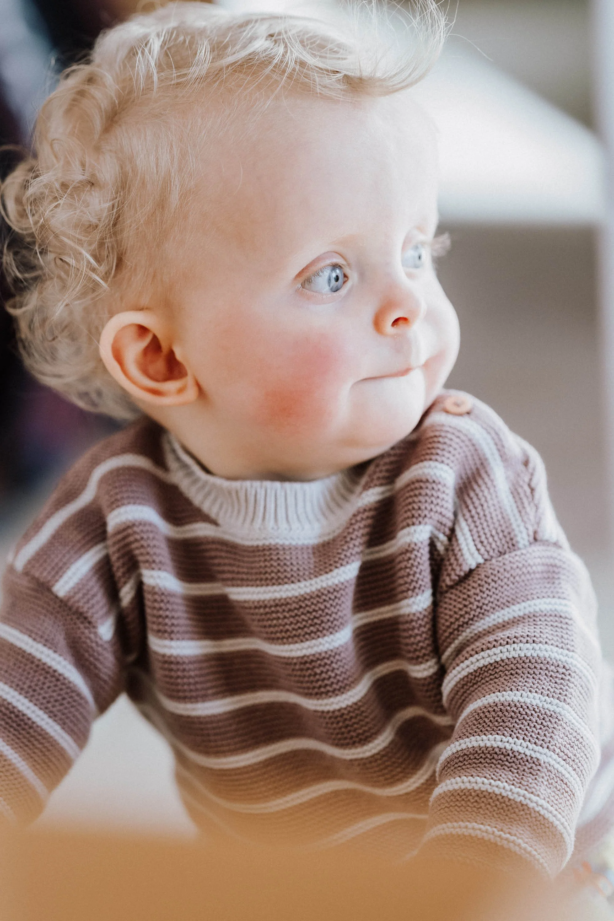 Ein kleiner Junge mit blonden, lockigen Haaren und blauen Augen trägt ein braunes und weiß gestreiftes Pullover.