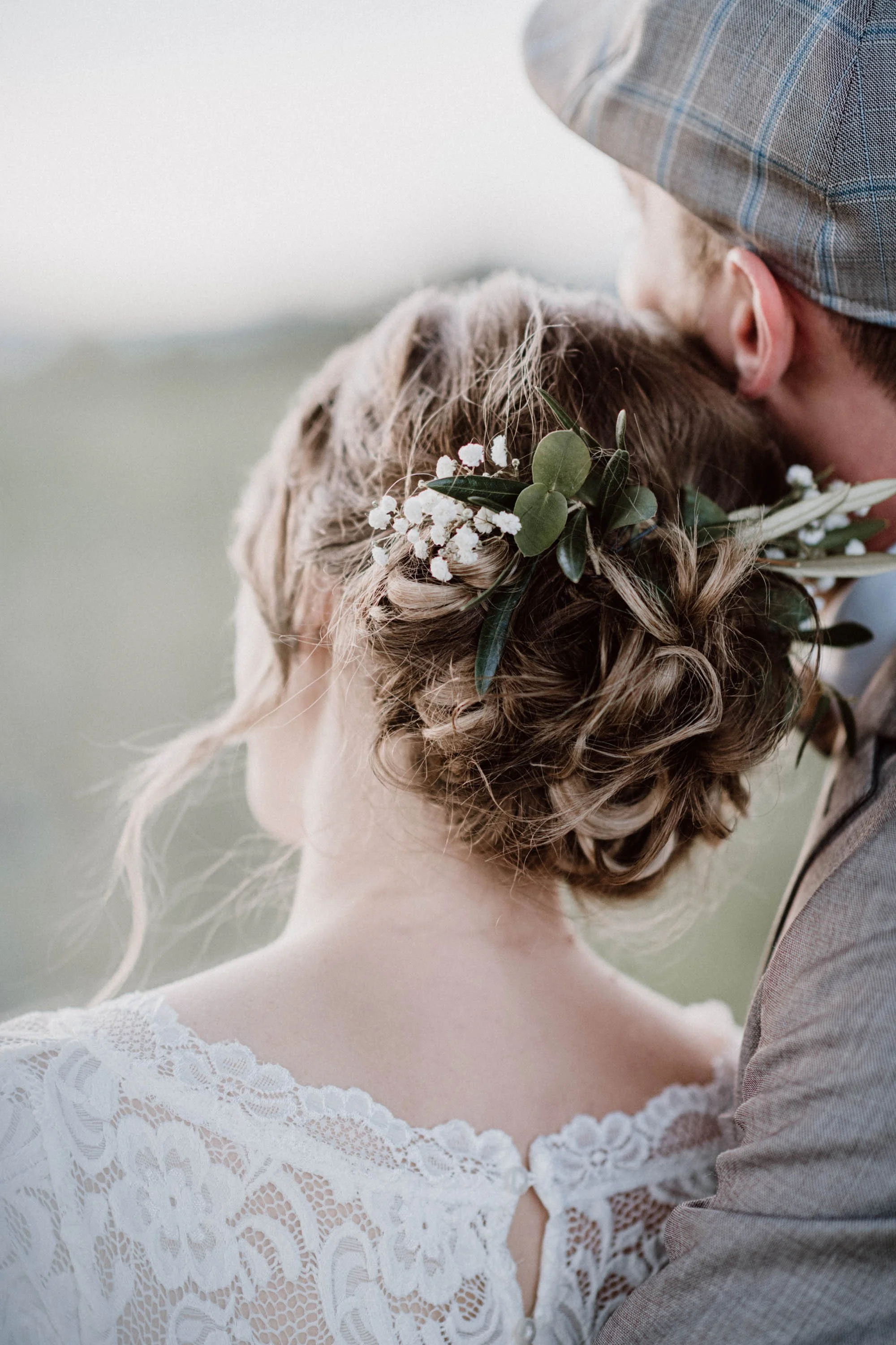 Nahaufnahme eines Brautpaares, das sich küssend umarmt, mit Fokus auf die Braut mit einer geflochtenen Frisur und Blumenakzent sowie den Bräutigam in einem grauen Anzug und Kappe.
