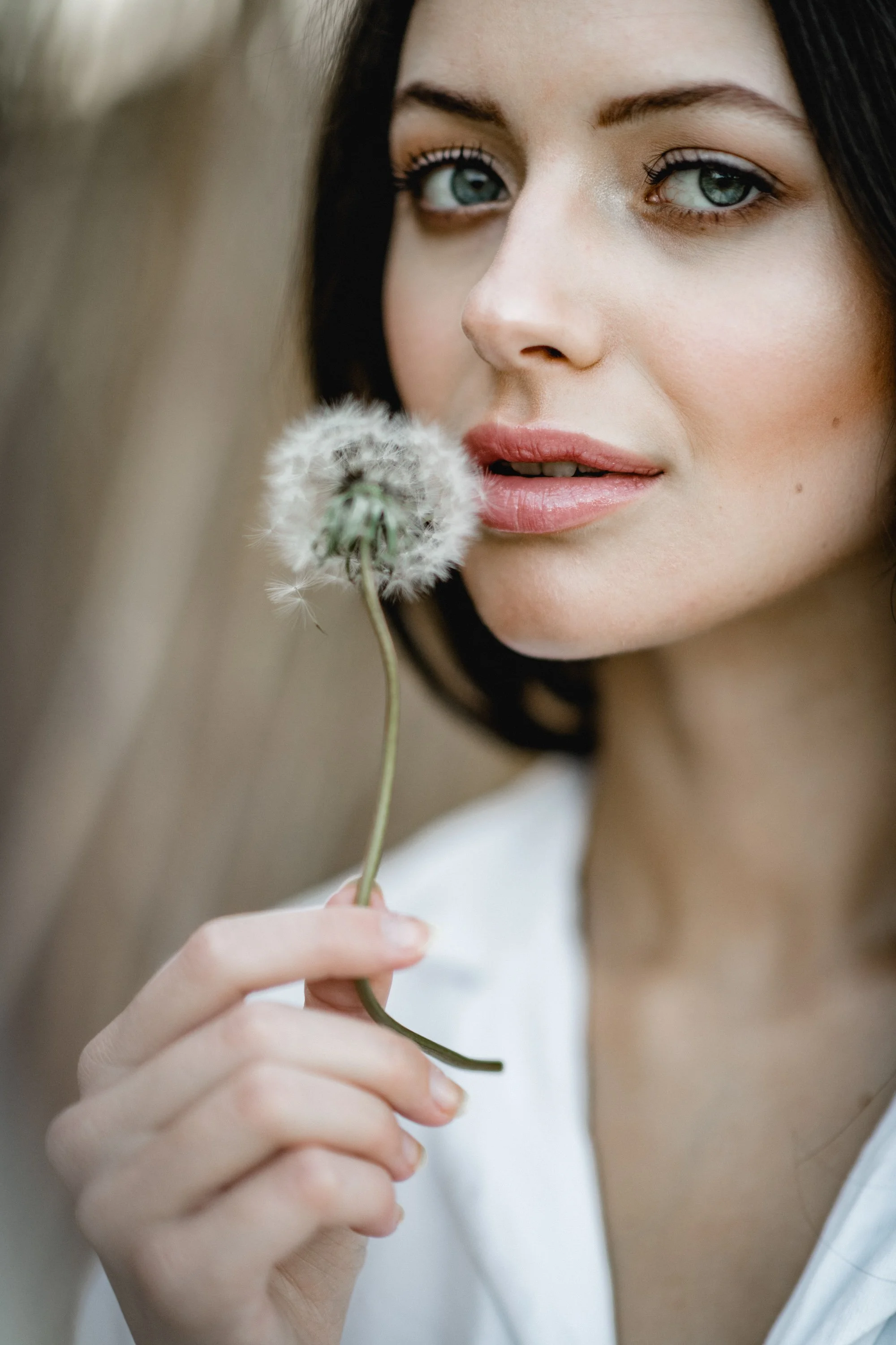 Nahaufnahme eines Mädchens mit dunklen Haaren, blauen Augen und naturlangem Make-up, das eine Pusteblume hält.