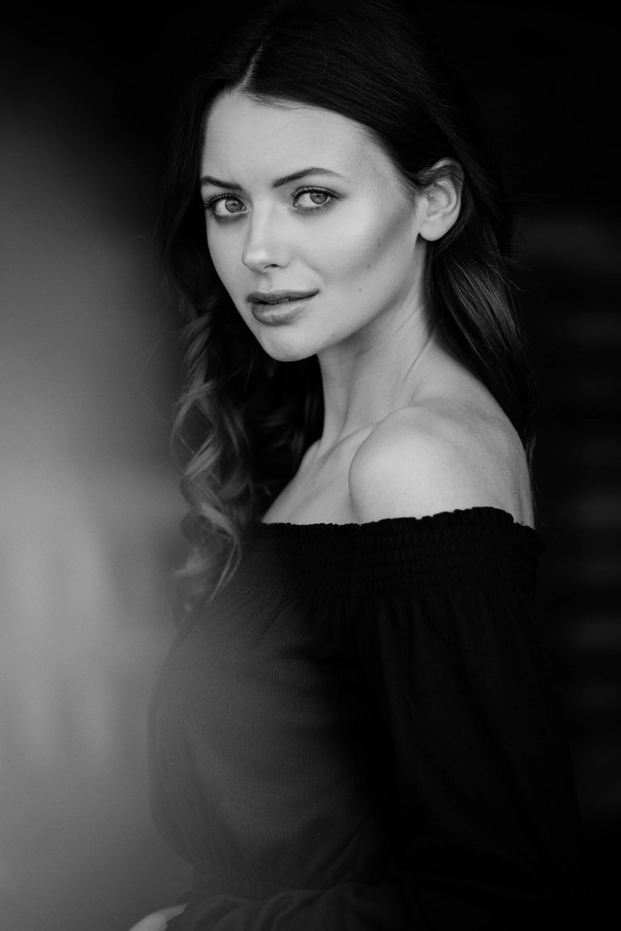 Schwarz-weiß Porträt einer jungen Frau mit lang, gewelltem Haar, die eine schwarze Off-Shoulder-Bluse trägt.