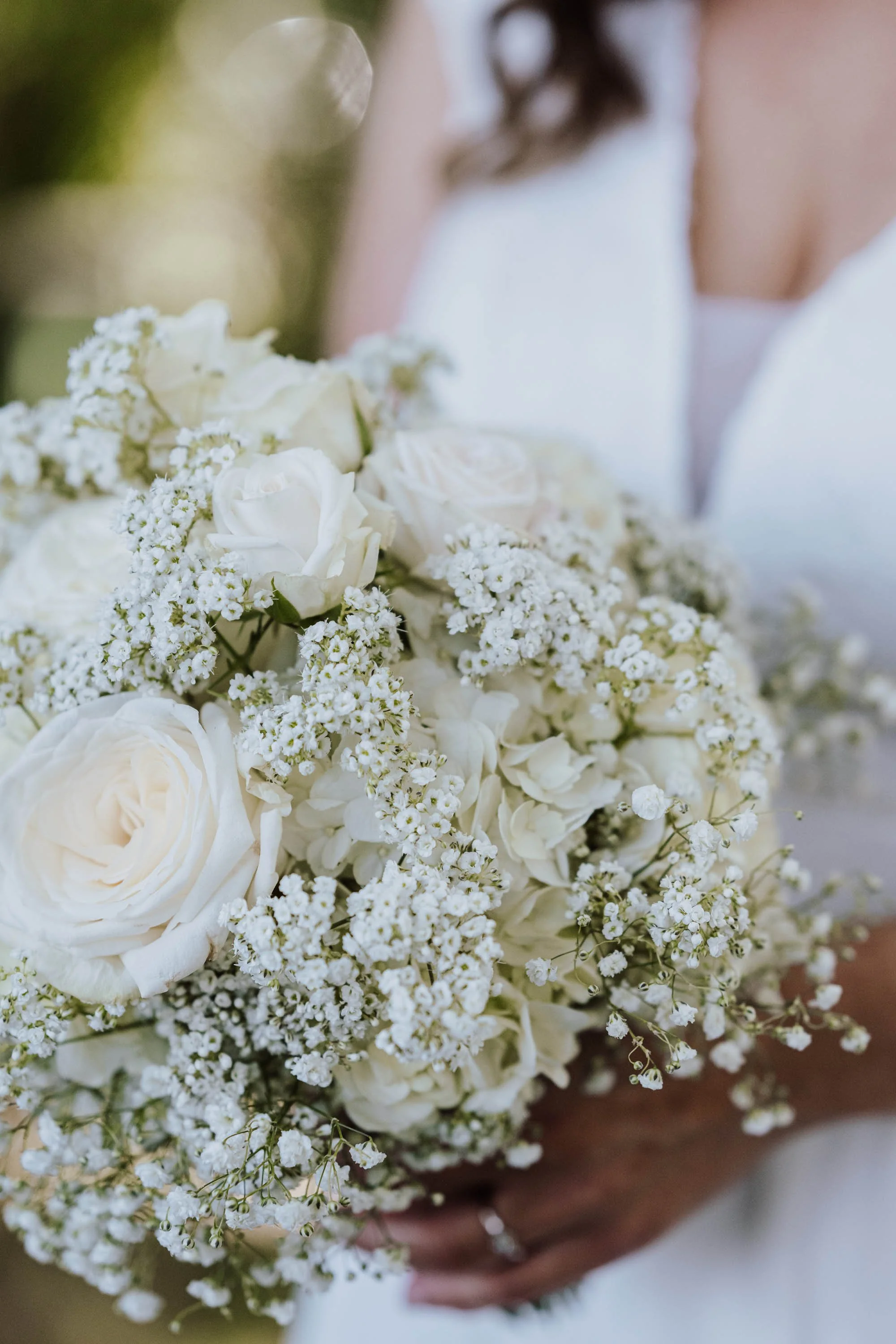 Eine Hand hält einen eleganten Brautstrauß aus weißen Rosen, kleinen weißen Blümchen und Schleierkraut, bei einer Hochzeit.
