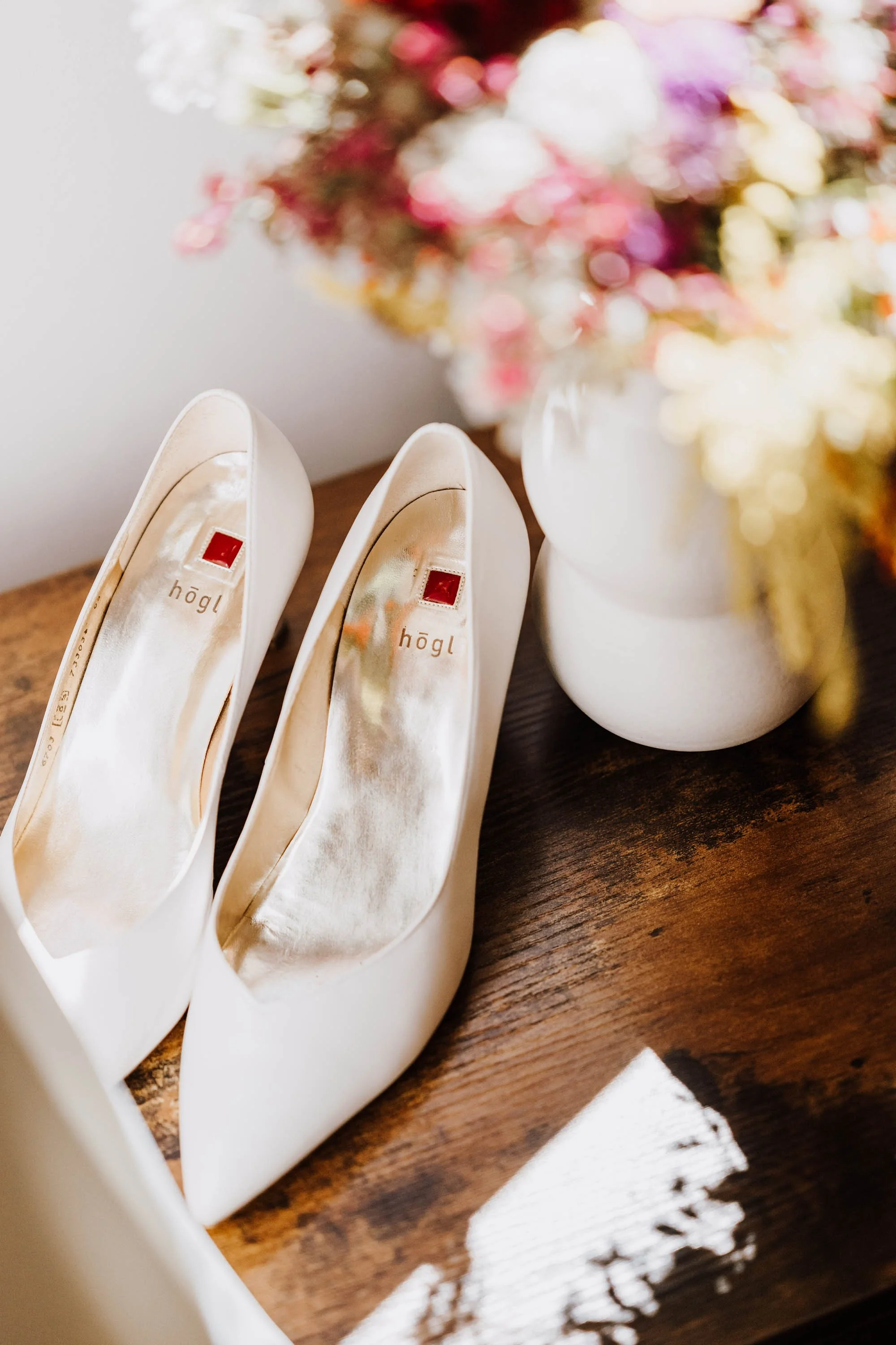 Weiße High Heels neben einem Blumenstrauß in einer weißen Vase auf einem Holztablett