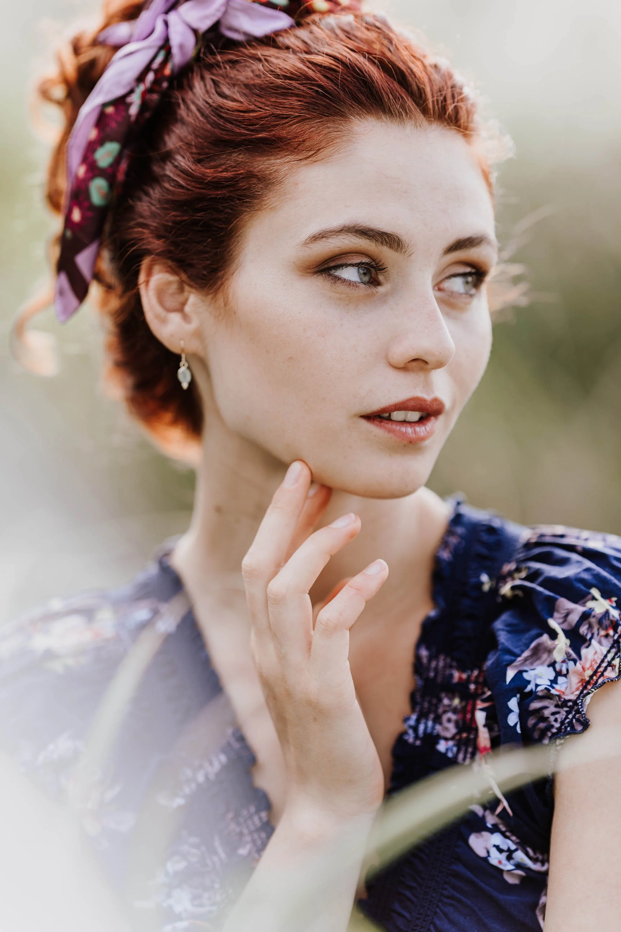 Ein Porträt einer jungen Frau mit roten Haaren, die einen Schal im Haar trägt, trägt ein dunkelblaues, florales Kleid im Freien.