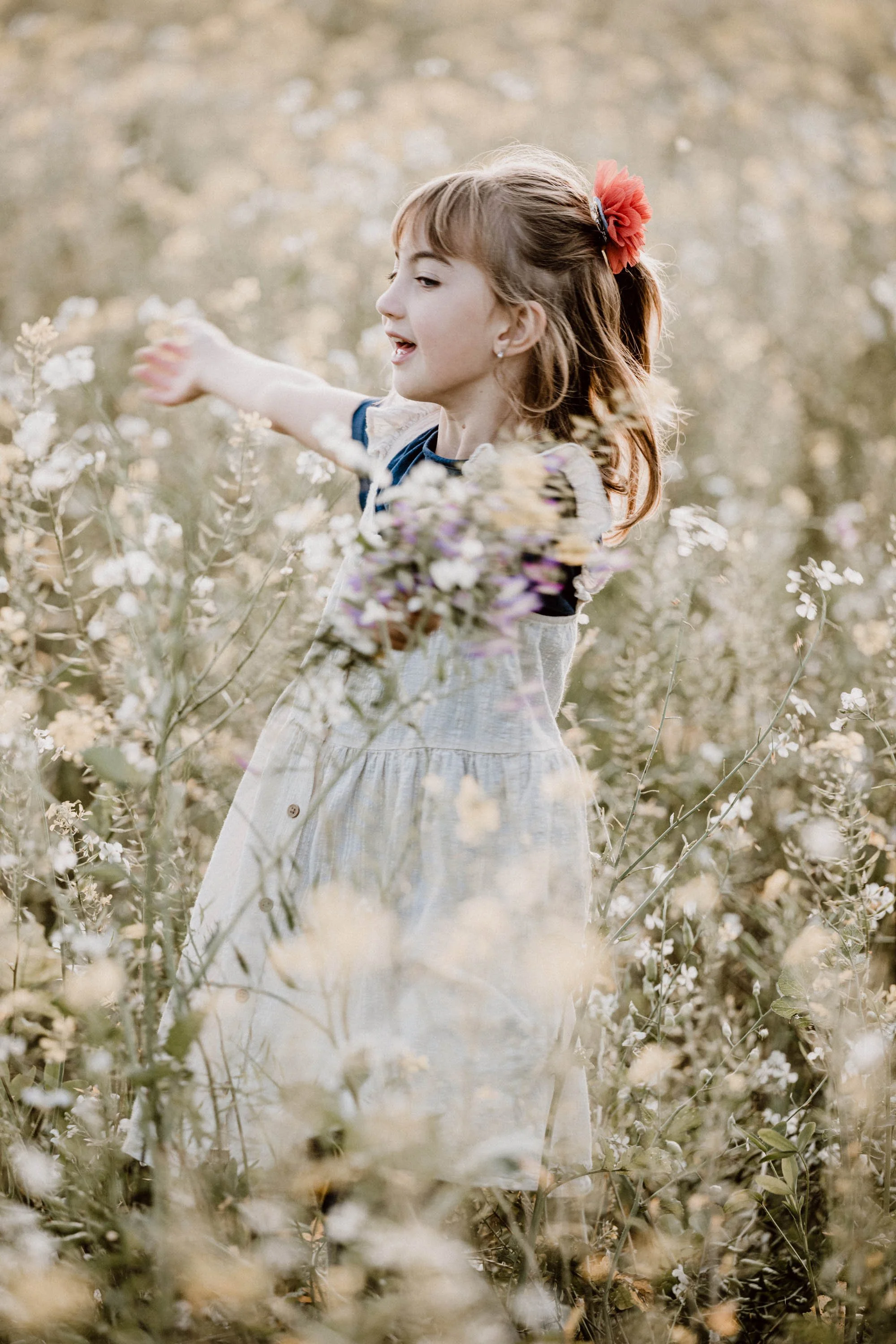 Ein Mädchen mit braunem Haar, das eine rote Haarspange trägt, steht in einem blühenden Blumenfeld und streckt einen Arm aus.
