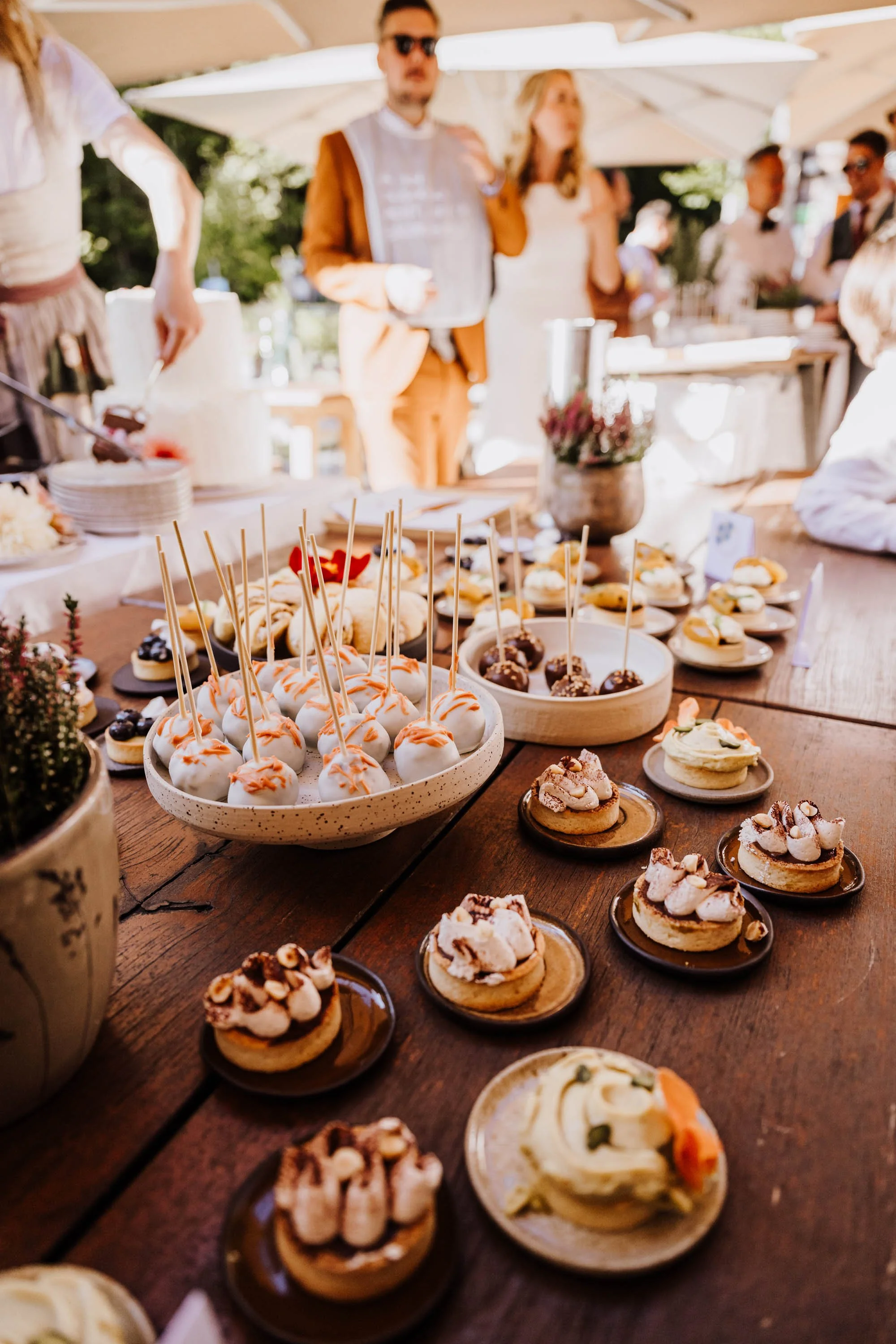 Verschiedene kleine Desserts und Kuchen auf einem Holztisch bei einer Feier im Freien, im Hintergrund stehen Menschen, einige unter einem Sonnenschirm.