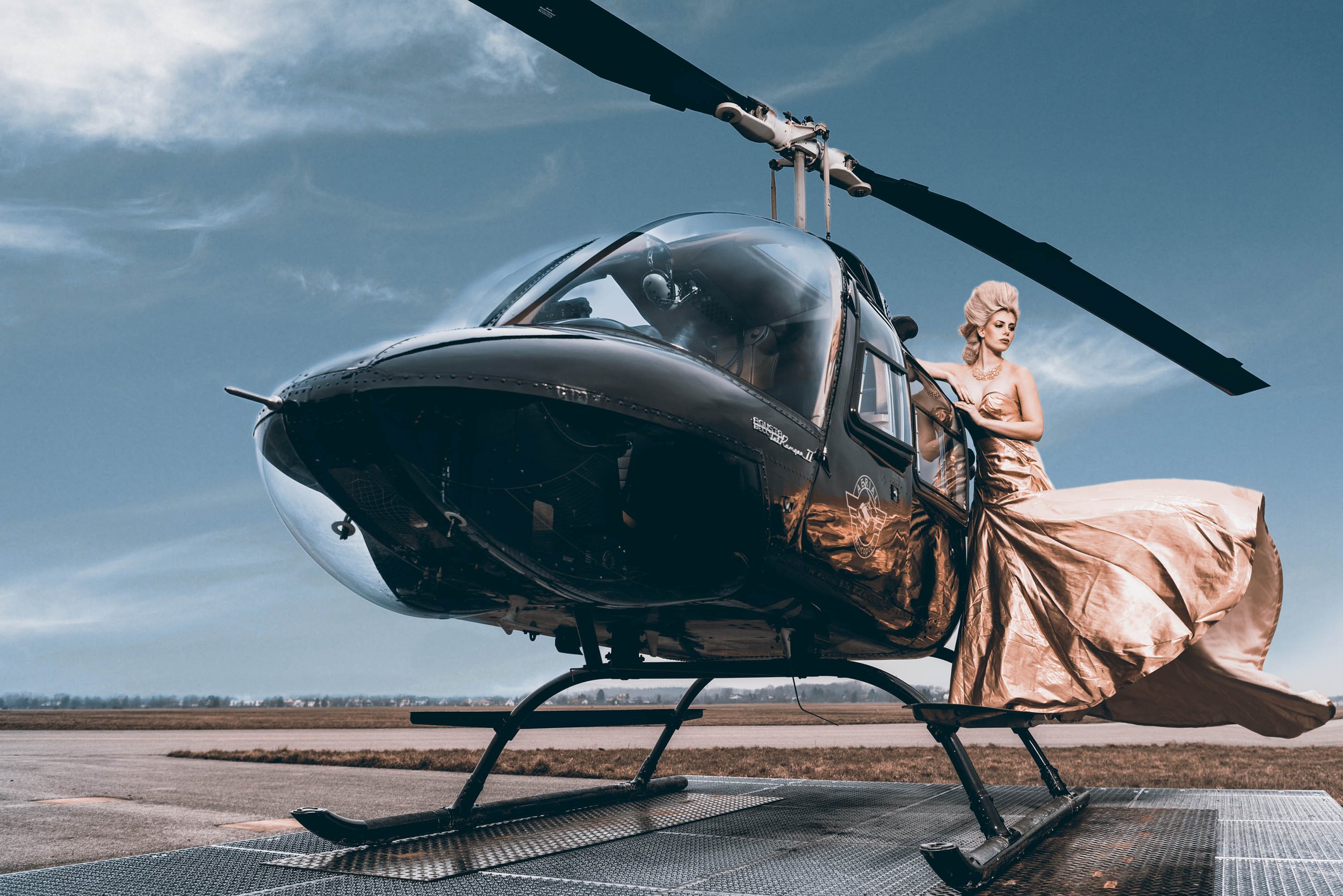Frau in goldener Abendkleidung steht neben einem schwarzen Hubschrauber auf einem Flugfeld bei bewölktem Himmel.