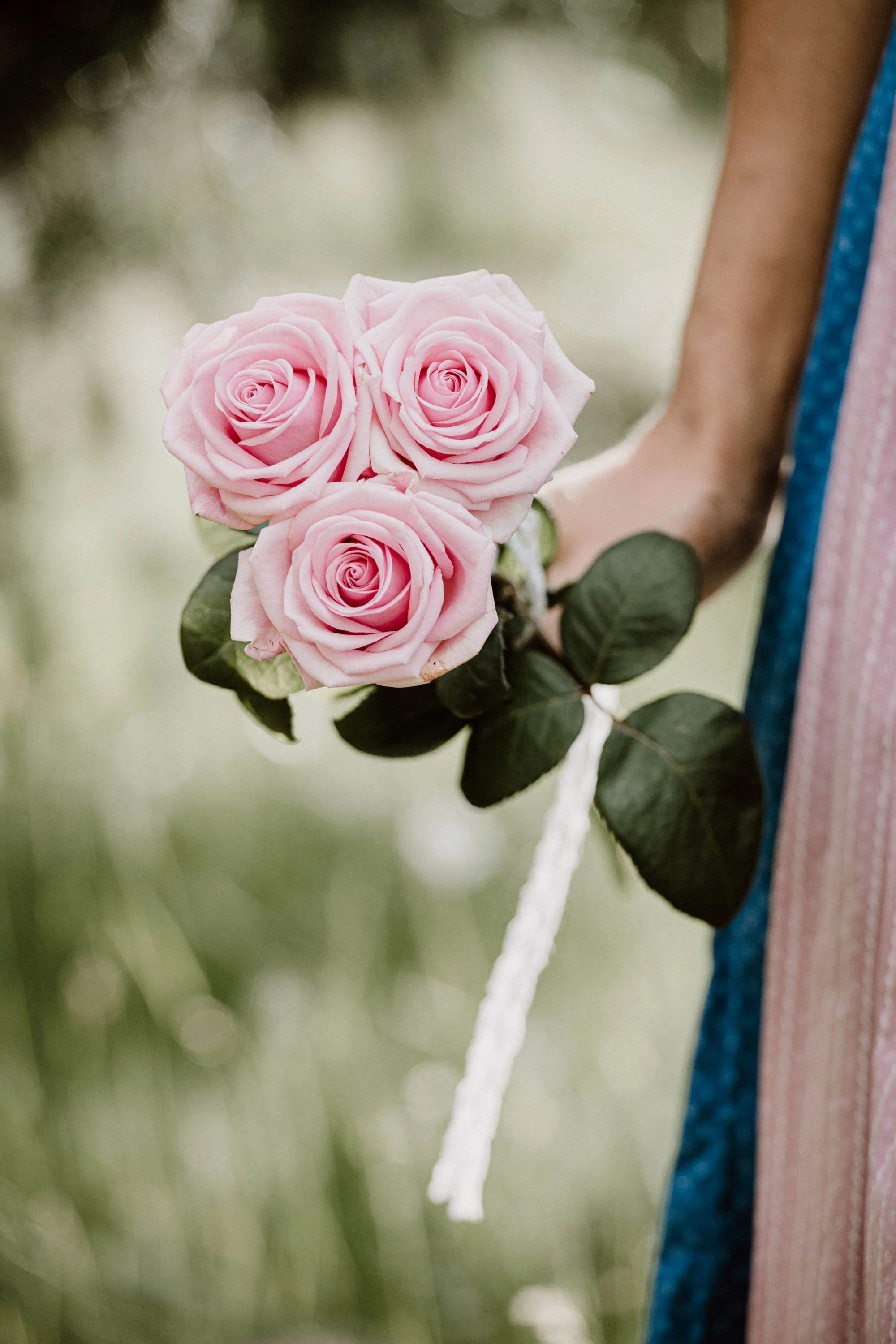 Hand hält einen Strauß aus drei pinken Rosen mit grünen Blättern bei einem natürlichen Hintergrund.