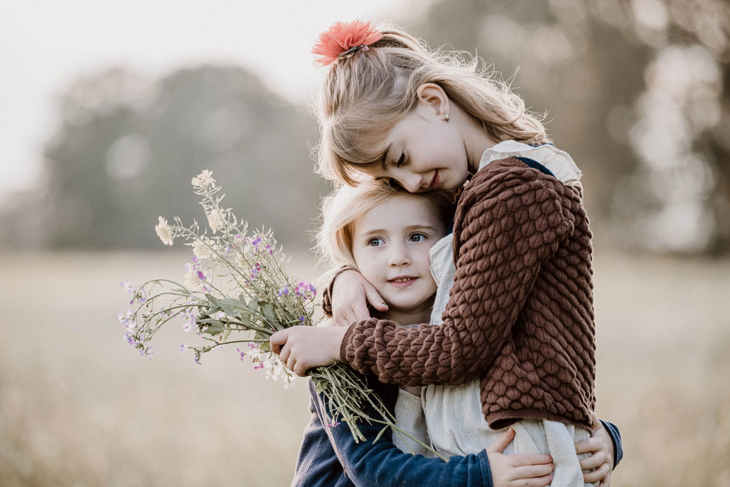 Zwei Mädchen umarmen sich auf einer Wiese, eines hält einen Blumenstrauß, im Hintergrund unscharfer Himmel und Bäume.