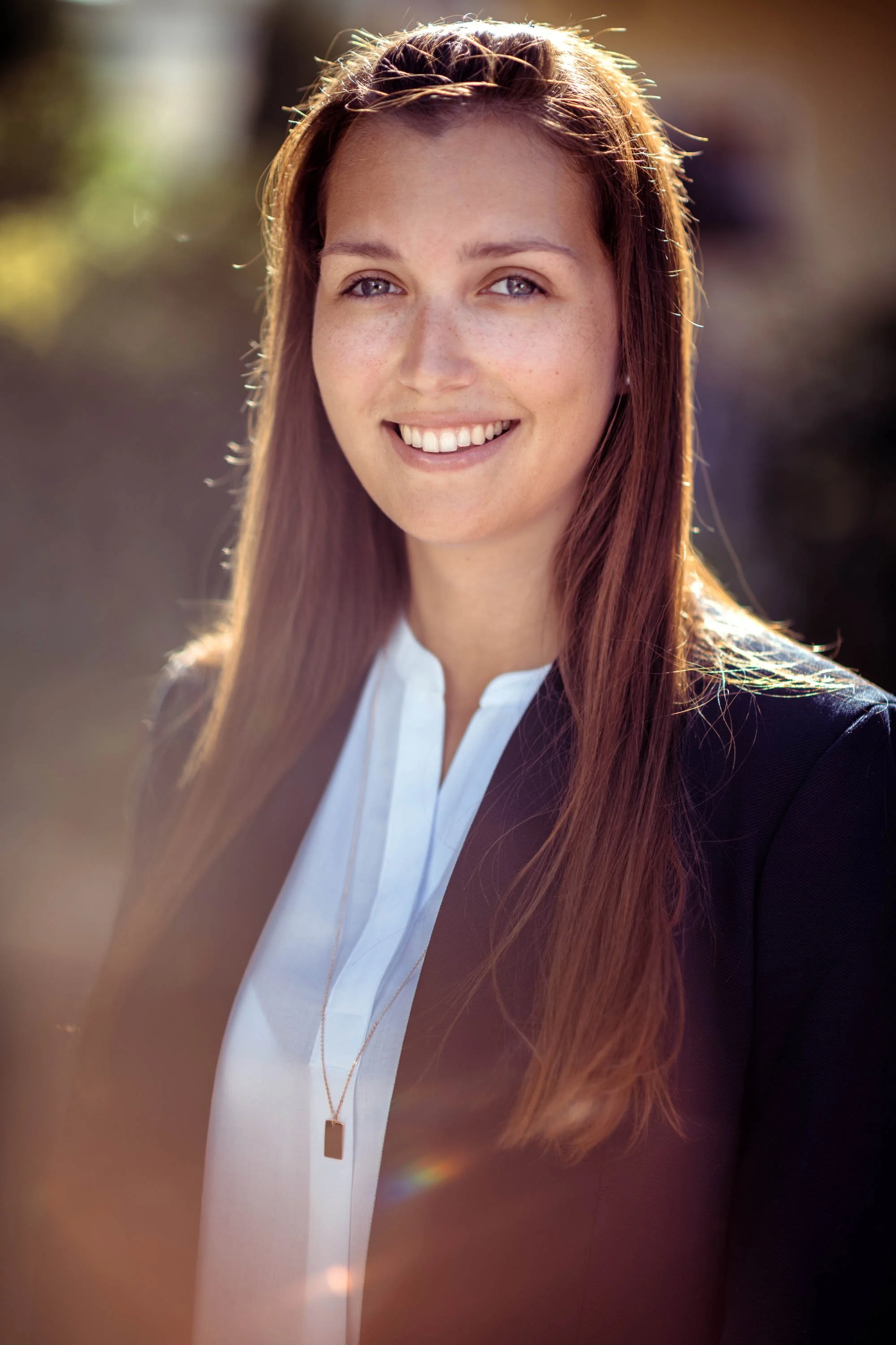 Junge Frau mit langen braunen Haaren, weißem Hemd und dunklem Blazer, lächelnd im Sonnenlicht, outdoor.