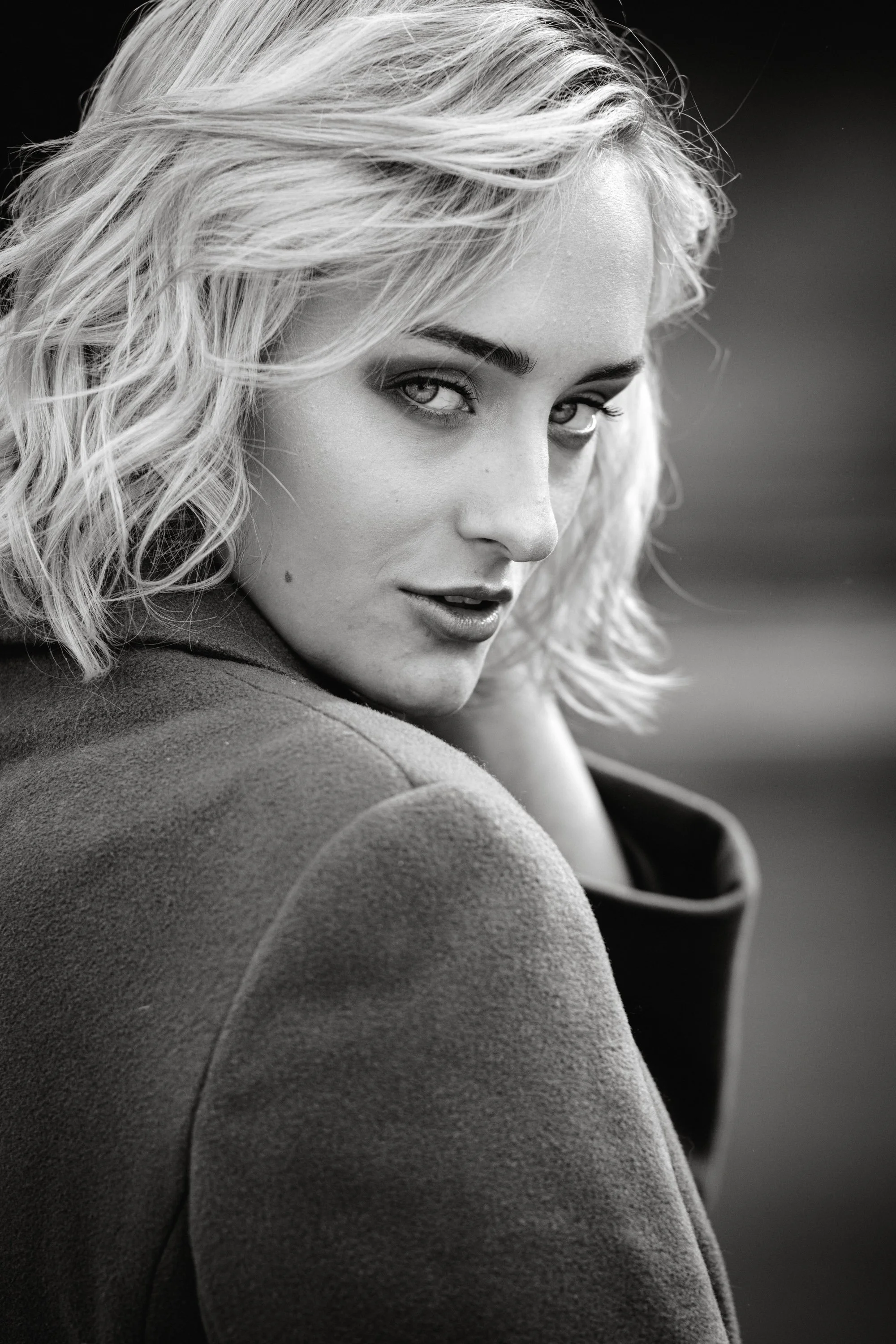 Schwarz-weiß-Porträt einer jungen Frau mit lockigem, blonden Haar, seriösem Ausdruck, trägt eine dunkle Jacke.