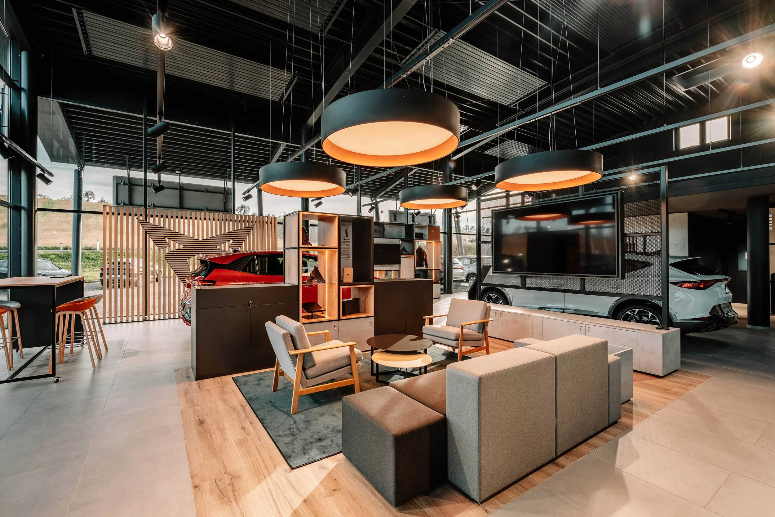 Ein modernes Autohaus mit weißen und roten Autos, Lounge-Bereich mit Sofas, Sesseln und TV, große Fenster, moderne Beleuchtung, Holzdielen und Steinkacheln im Boden.