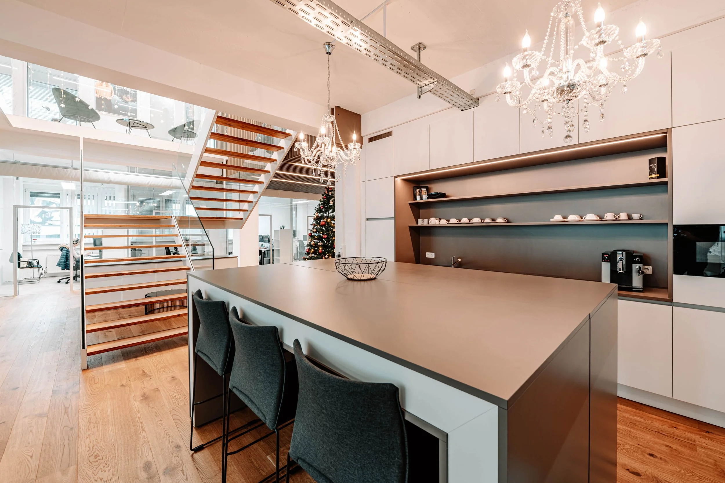 Moderne Küche mit zentralem Esstisch, schwarzen Stühlen, offenem Regal mit Geschirr, einem Kaffeebereich, Kronleuchtern und Weihnachtsbaum im Hintergrund.