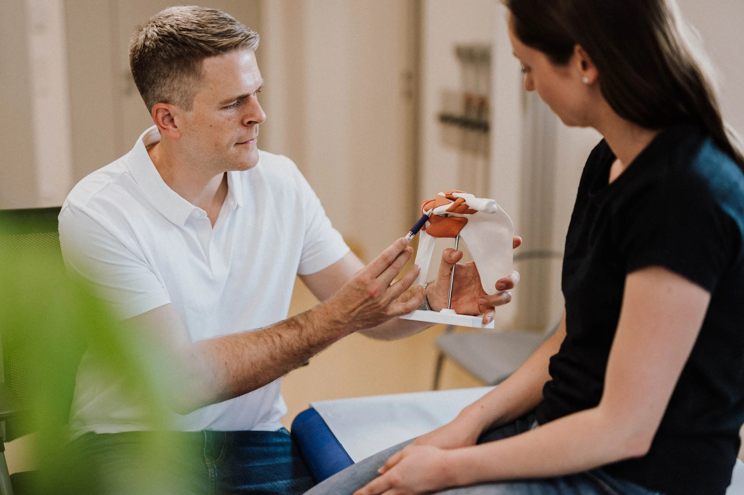 Arzt zeigt einer Patientin ein Modell des Schultergelenks während einer Beratung im medizinischen Wartezimmer.