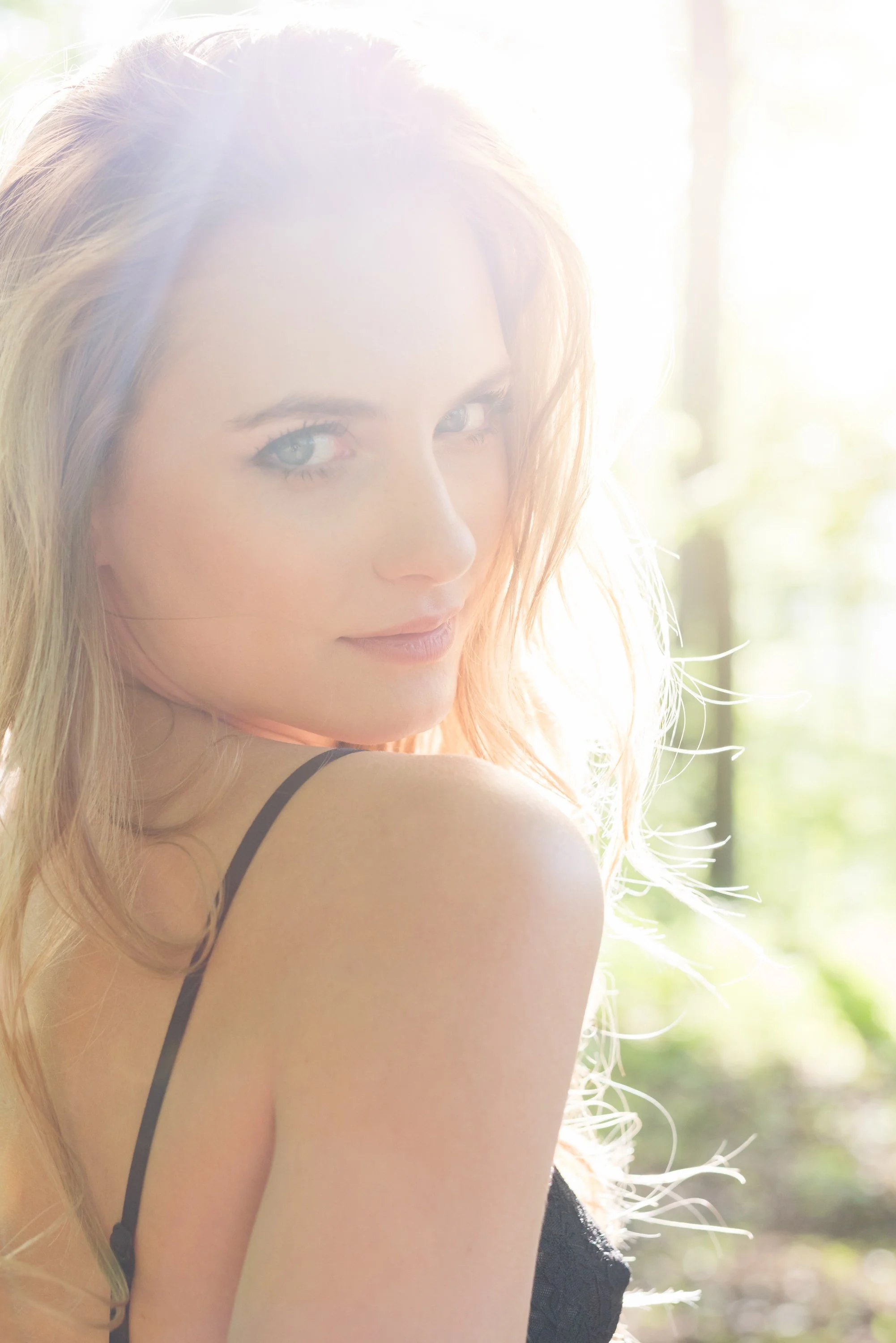 Nahaufnahme eines lachenden Mädchens mit langen blonden Haaren, das im Sonnenlicht steht, im Hintergrund unscharfer Wald.