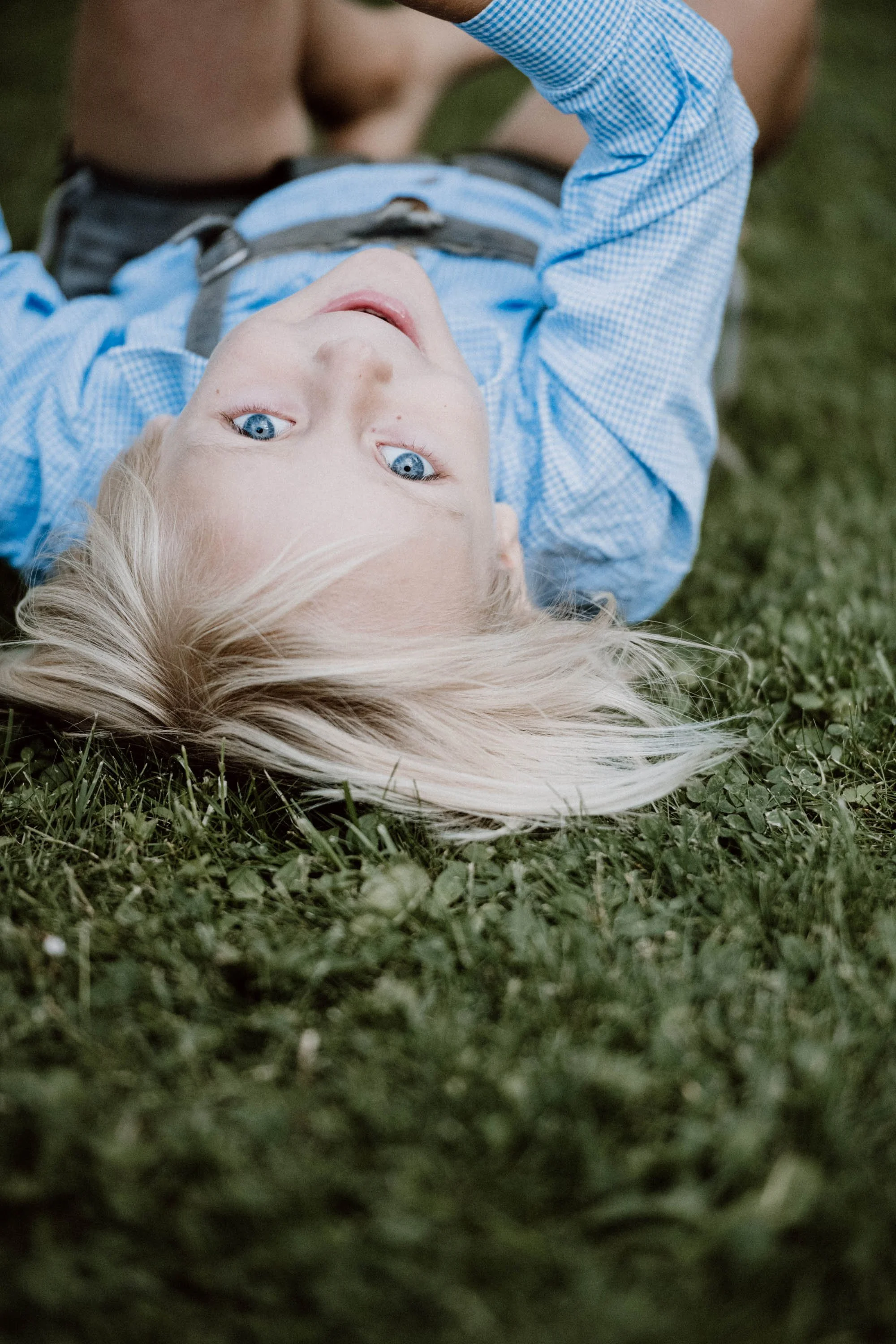 Ein Junge liegt auf dem Rasen, schaut direkt in die Kamera, hat blonde Haare, trägt blaue Kleidung, im Hintergrund sind andere Personen zu sehen.