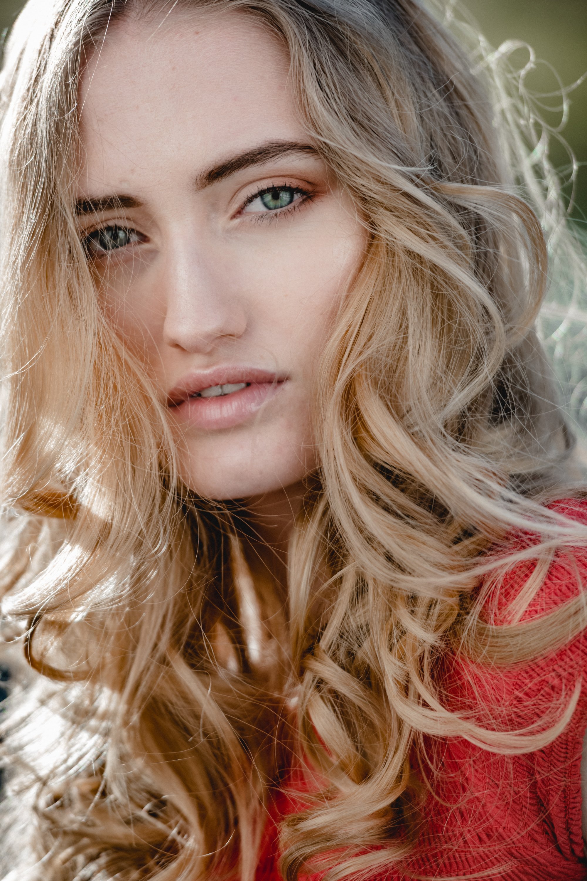 Nahaufnahme eines jungen Mädchens mit blonden, lockigen Haaren und blauen Augen, trägt ein rotes Oberteil, im Freien bei sonnigem Licht.