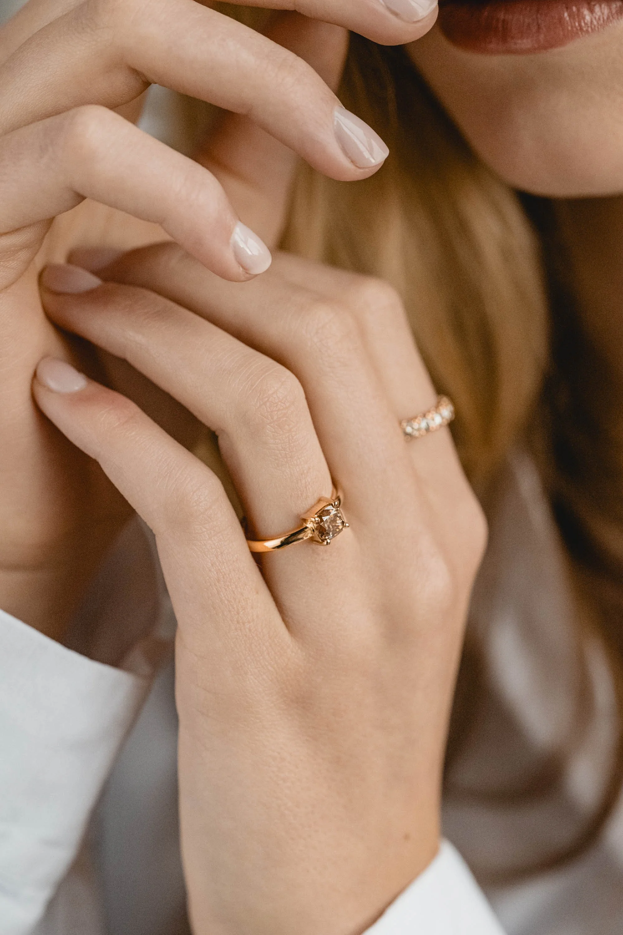 Nahaufnahme einer Hand mit einem Goldring, der einen großen, klaren Edelstein trägt, und einer anderen Hand mit einem schmalen Bandring mit kleinen Edelsteinen. Die Hände sind zart und gepflegt.