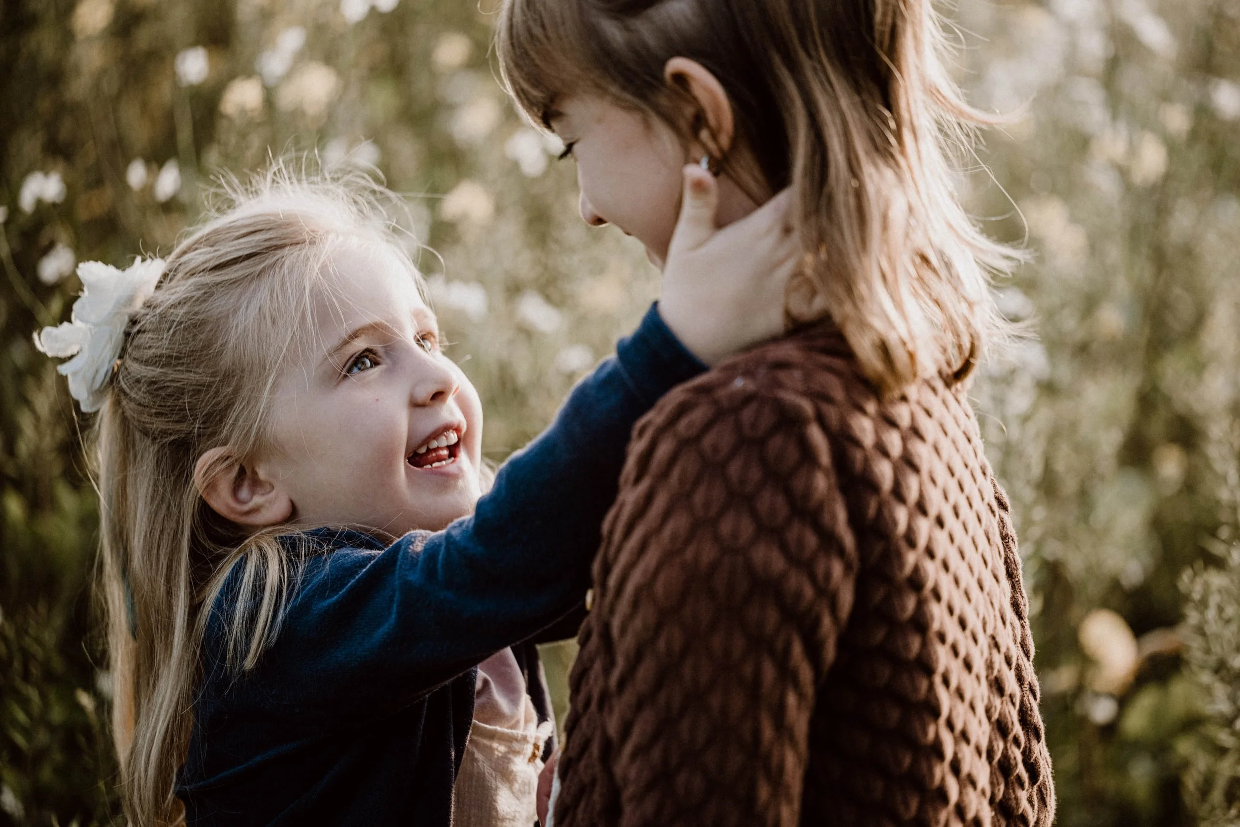 Eine junge Frau und ein kleines Mädchen, das liebevoll an ihrem Hals zieht, sitzen im Freien vor einem blühenden Busch im Sonnenlicht.