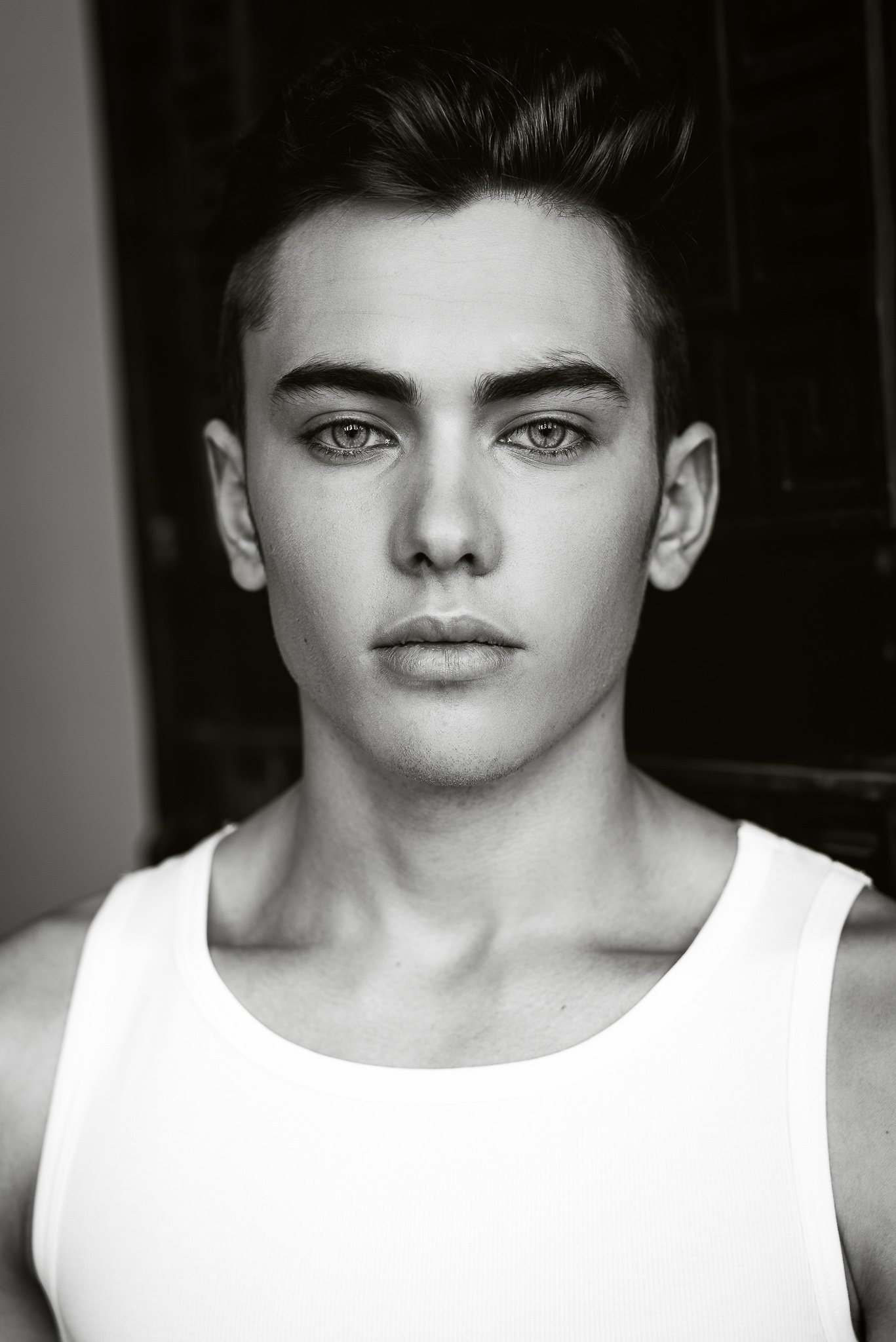 Schwarz-Weiß-Porträt einer jungen Person mit kurzer, dunkel gelockerter Frisur, trägt ein weißes Tanktop, Blick gerichtet in die Kamera, expressionistische Mimik.