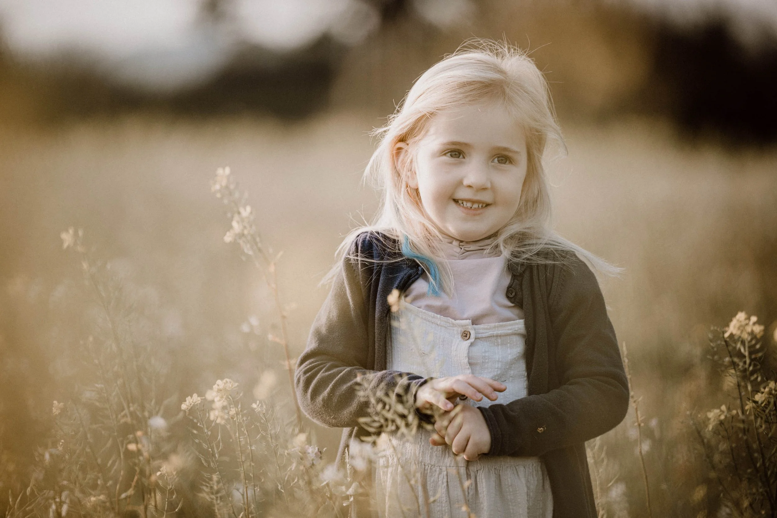 Ein junges Mädchen mit blonden Haaren, das in einem Feld mit Pflanzen steht, lächelt und in die Kamera schaut.