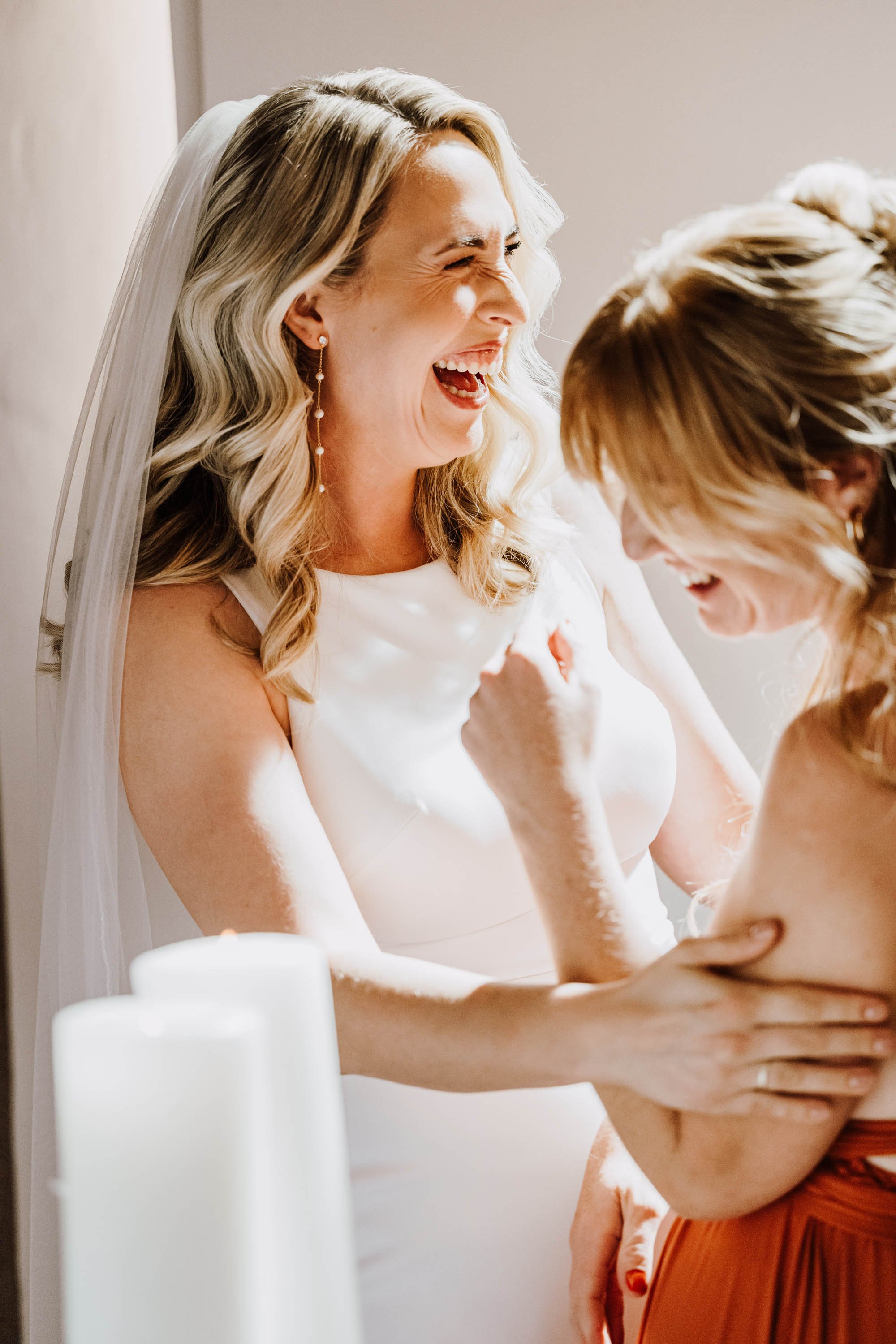 Zwei lachende Frauen, eine im Hochzeitskleid mit Schleier, die andere in einem roten Kleid, teilen einen fröhlichen Moment.