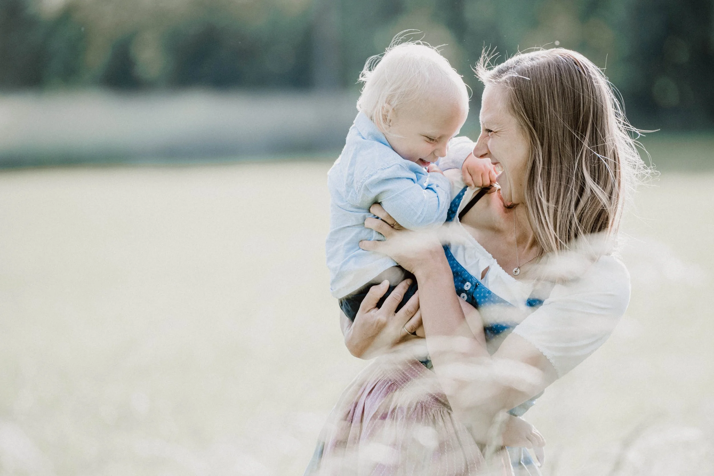 Eine lachende Frau hebt ein lachendes Kind in der Natur hoch, beide scheinen Spaß zu haben.