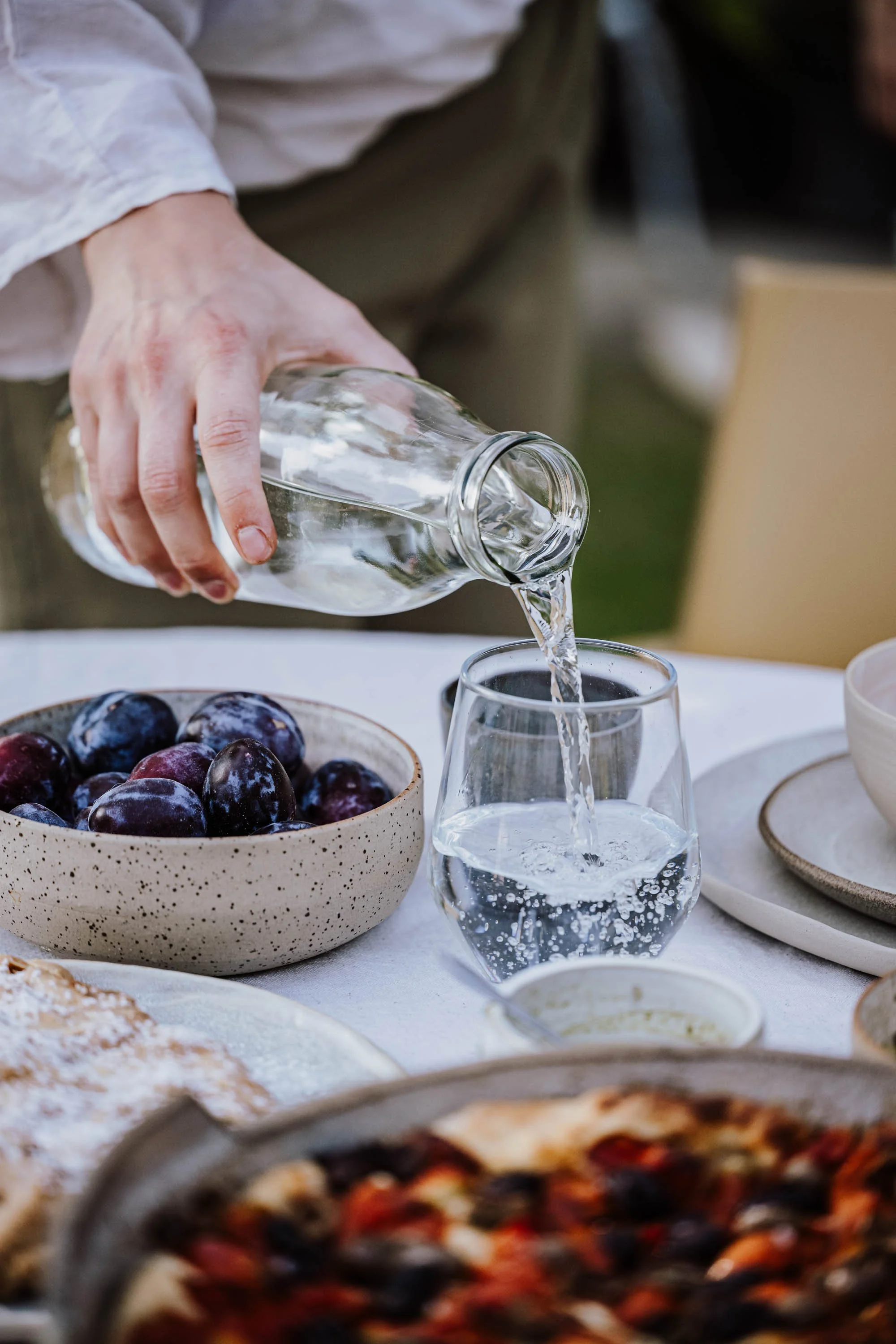 Hand, die Wasser in ein Glas gießt, auf einem gedeckten Tisch mit Obst und Essen, im Hintergrund Lebensmittel und Geschirr.