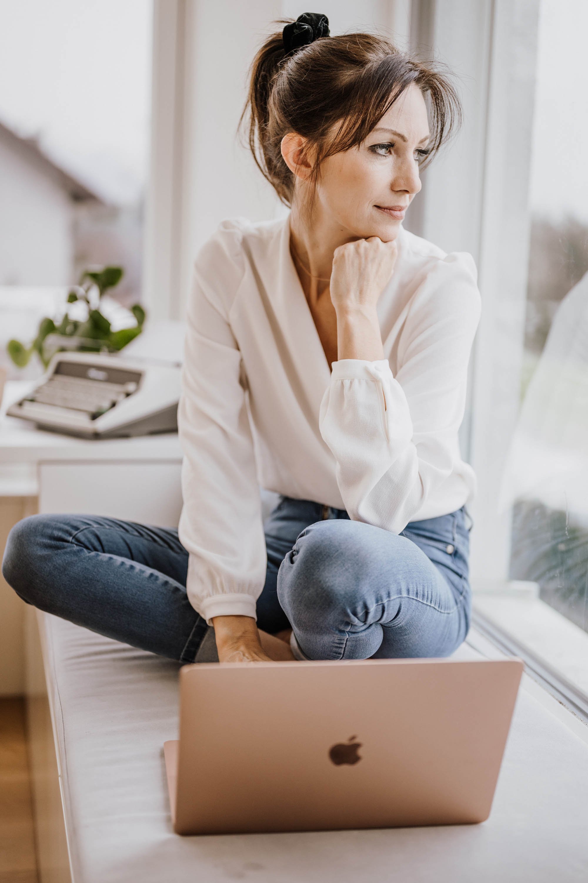 Frau sitzt nachdenklich auf einer Fensterbank mit einem offenen Laptop vor sich.