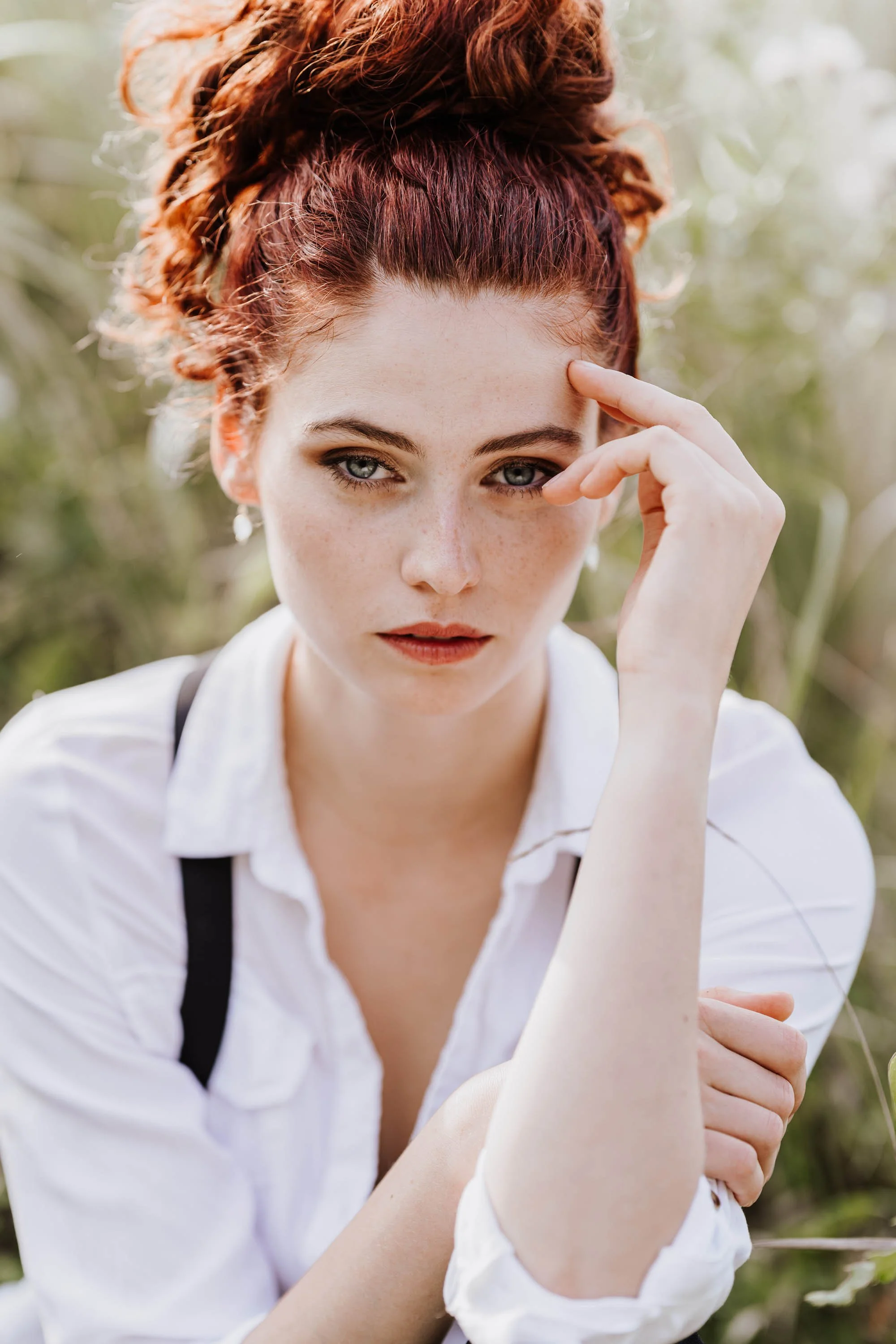 Junge Frau mit roten, lockigen Haaren und hellen Augen, trägt weiße Bluse, sitzt in der Natur, schaut nach vorne, während sie eine Hand an die Stirn legt.