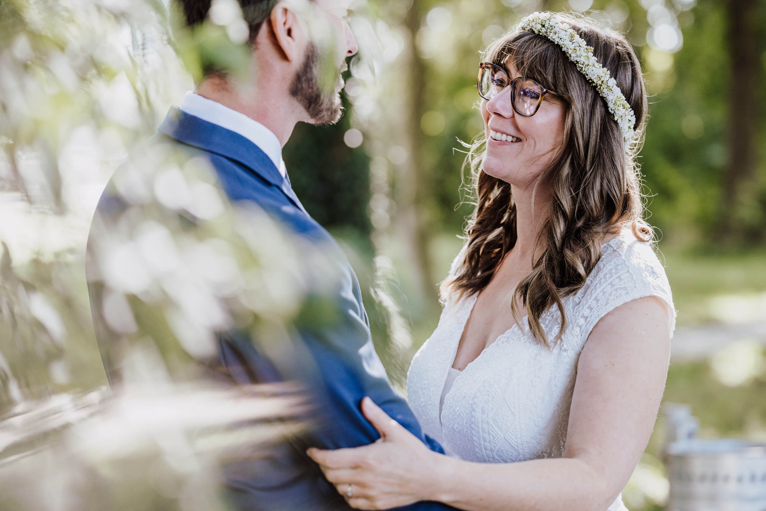 Ein Paar bei einer Hochzeit, die Frau trägt ein weißes Kleid mit Spitze und eine Blumenkrone im Haar, der Mann trägt einen blauen Anzug, sie lächeln sich im Freien an.