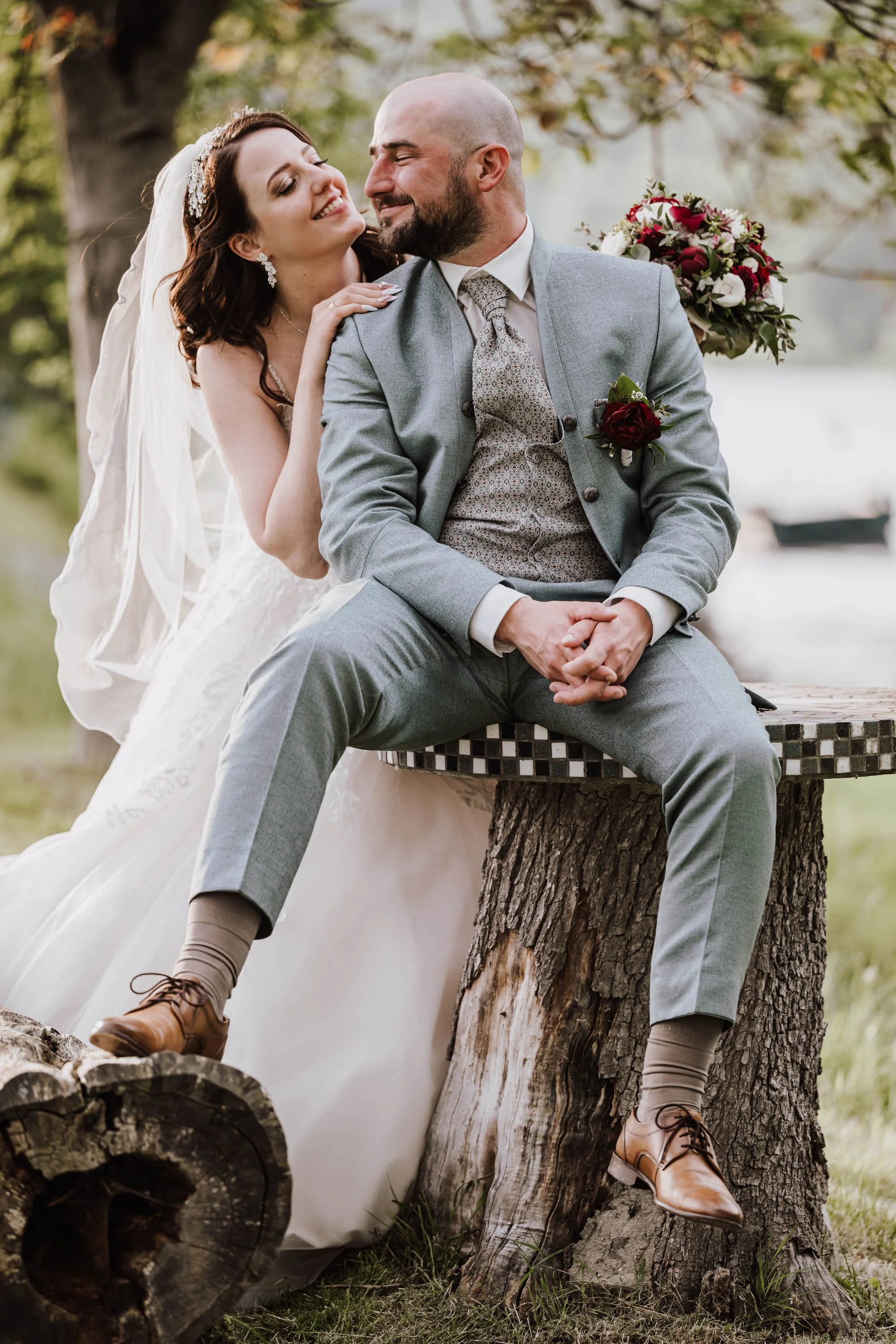 Braut und Bräutigam sitzen auf einem Baumstamm im Park, lachen und genießen den Moment, die Braut trägt ein weißes Hochzeitskleid und Schleier, der Bräutigam einen grauen Anzug, im Hintergrund Bäume und Wasser.