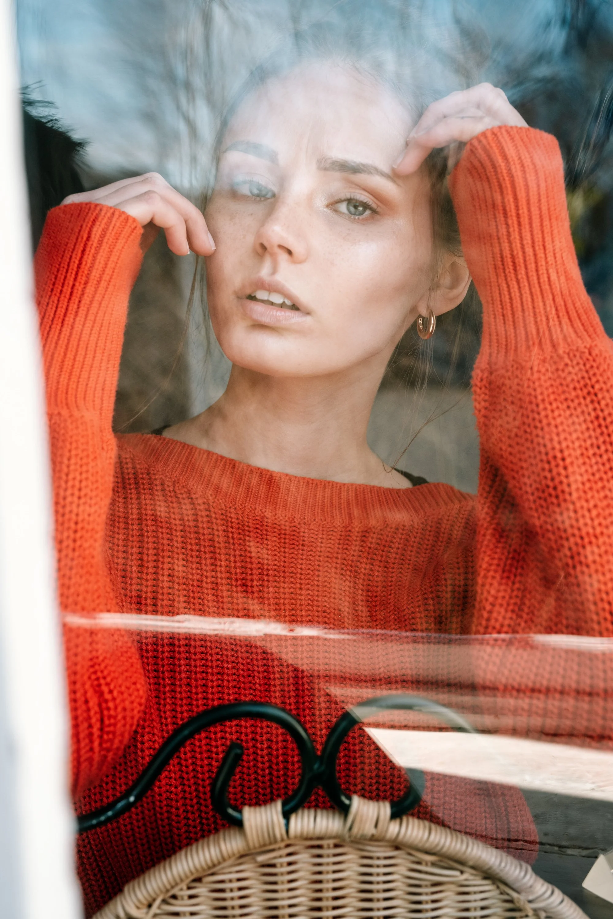 Junge Frau mit braunen Haaren und dezenten Make-up, trägt ein rotes Strickoberteil und eine kleine, goldene Ohrringe, blickt durch ein Fenster.