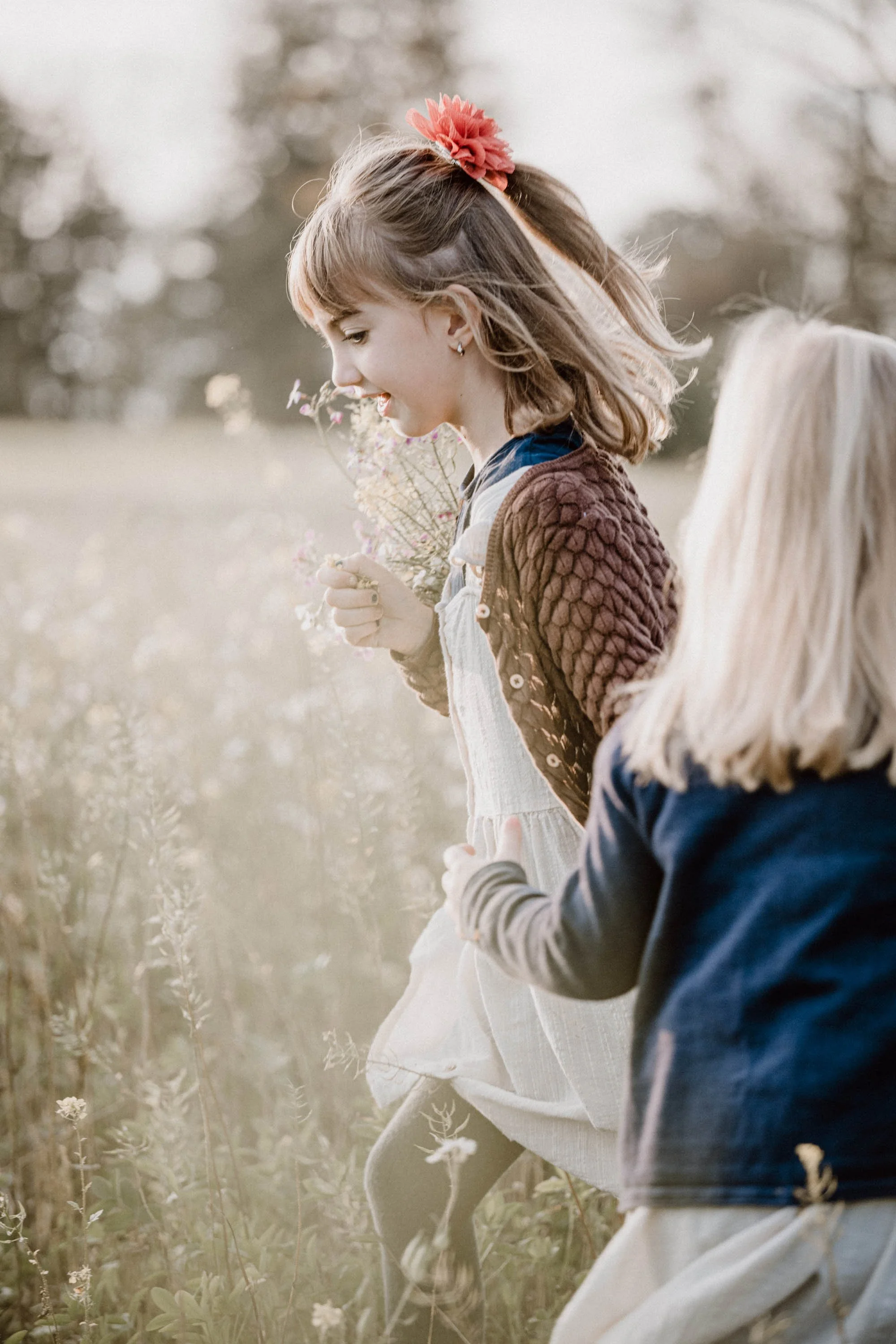 Zwei junge Mädchen in einem Feld, eines mit braunem Pullover und das andere mit blonder Haar und dunklem Jacke, sammeln Blumen.