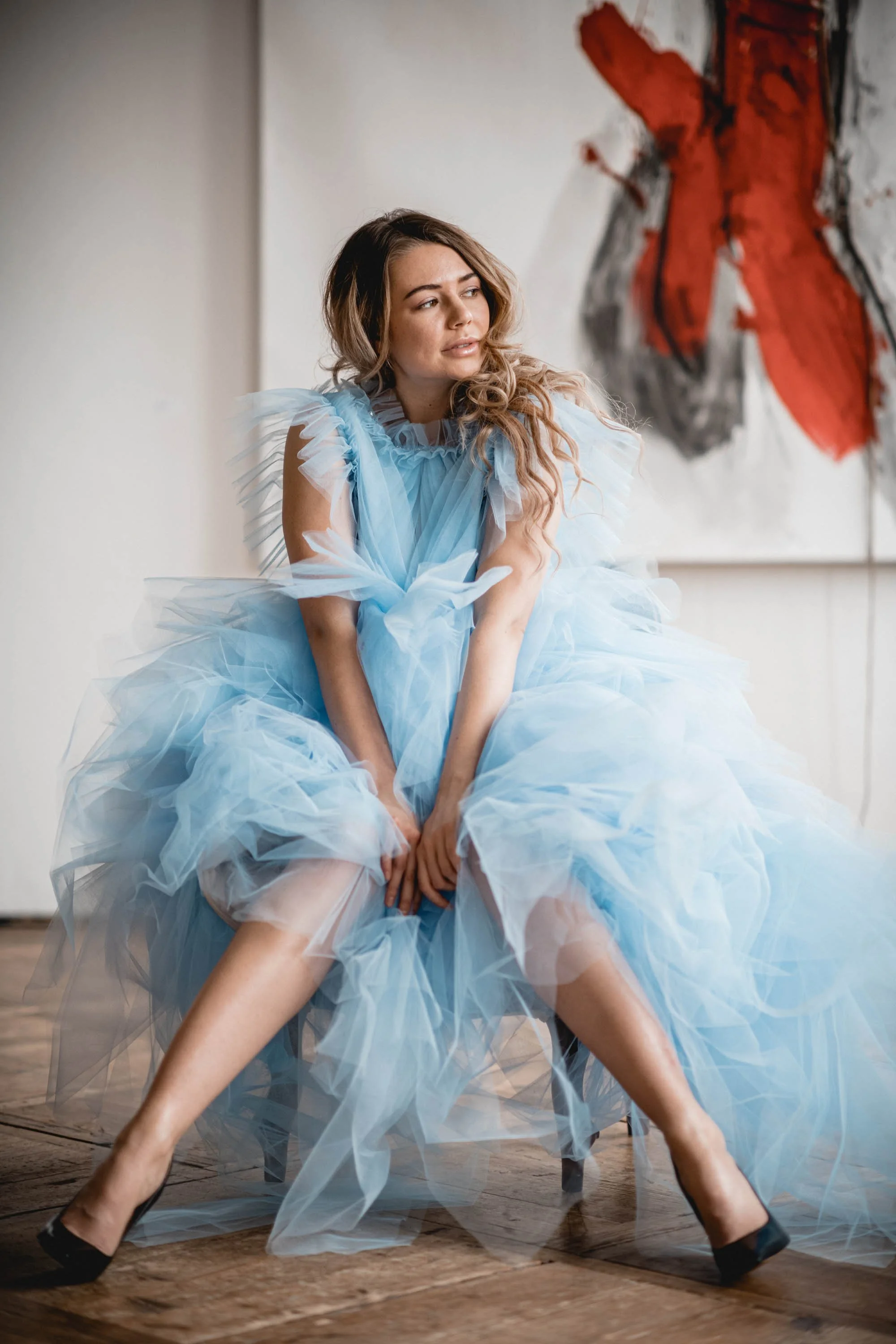 Frau in einem luftigen, blauen Tüllkleid sitzt auf einem Stuhl, im Hintergrund ein abstraktes Gemälde.