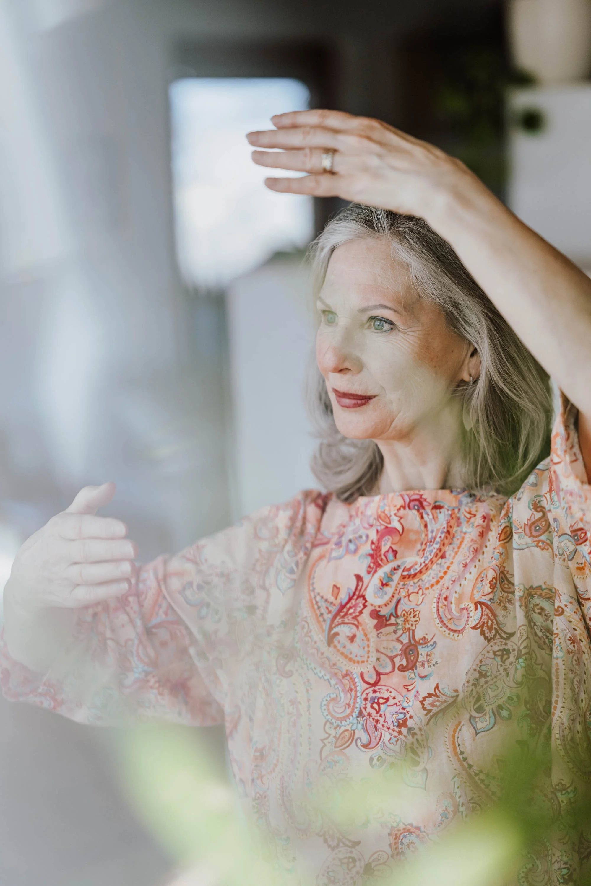 Eine ältere Frau macht eine Arm- und Handbewegung, vielleicht Yoga oder Tanzen, in einem hell erleuchteten Raum.