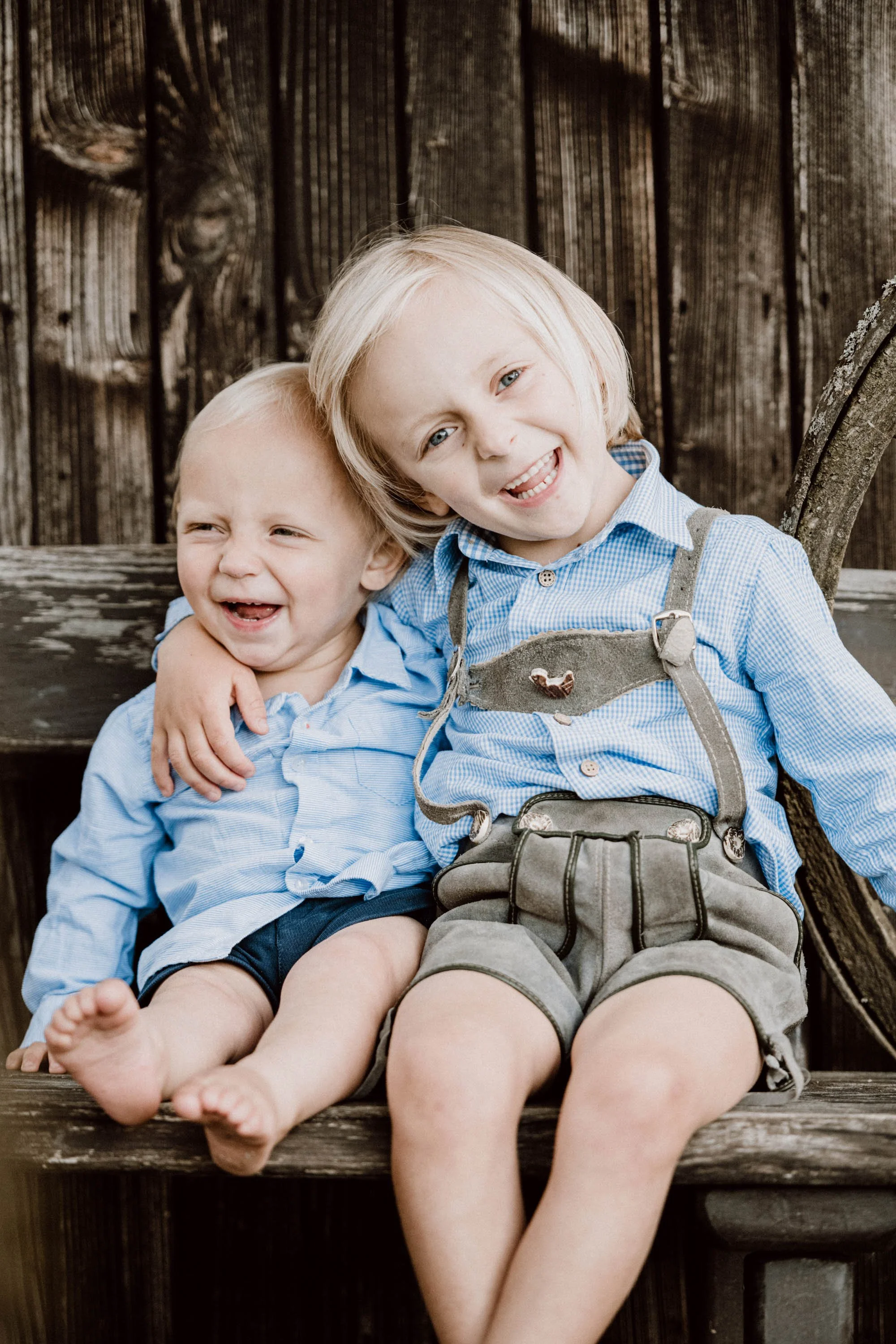 Zwei lachende junge Kinder, sitzend auf einer Holzbank, umarmend, vor einer Holzwand, tragen blaue Hemden.