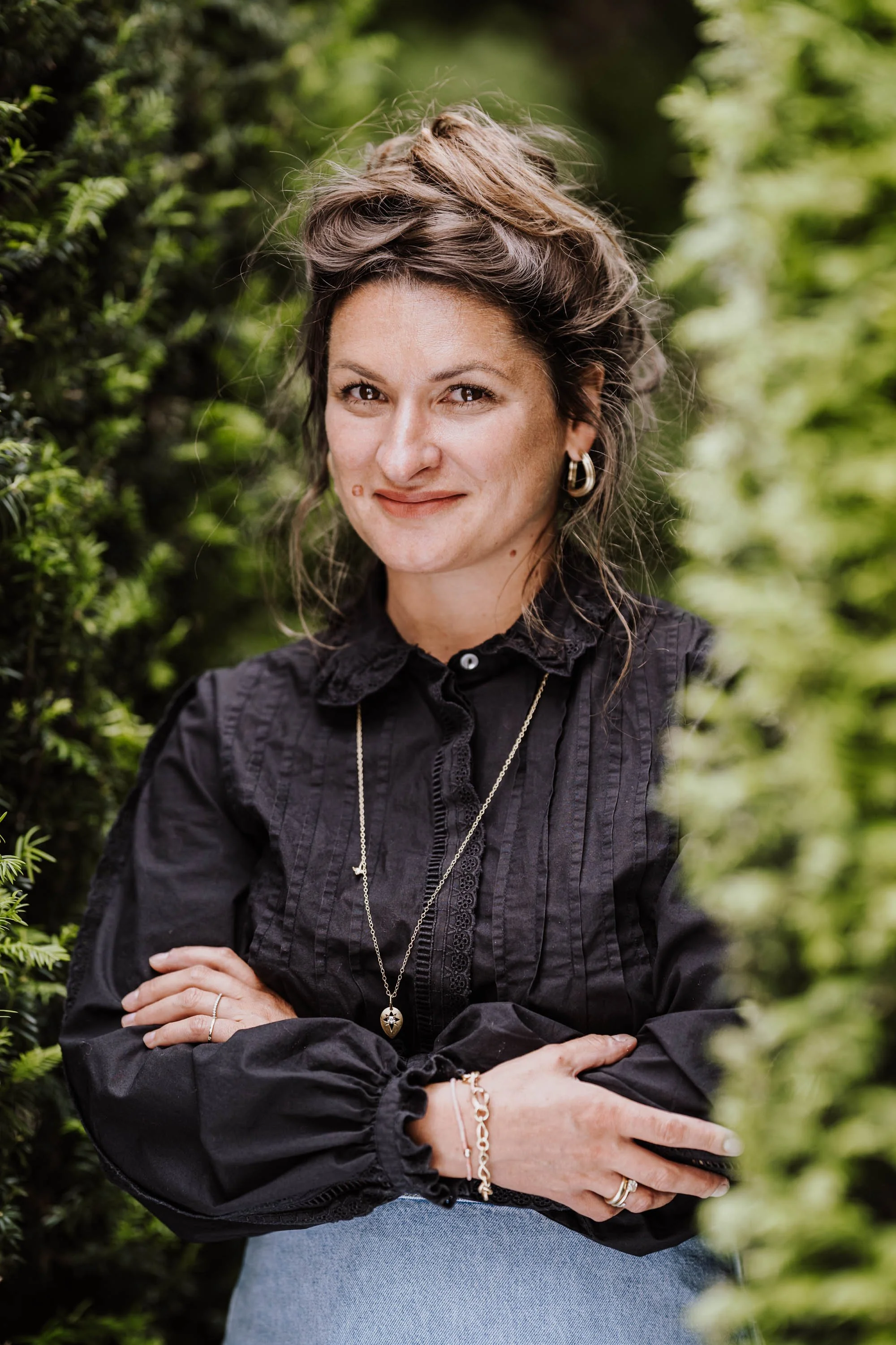 Porträt einer Frau mit dunklem Hemd, die zwischen grünen Büschen steht.