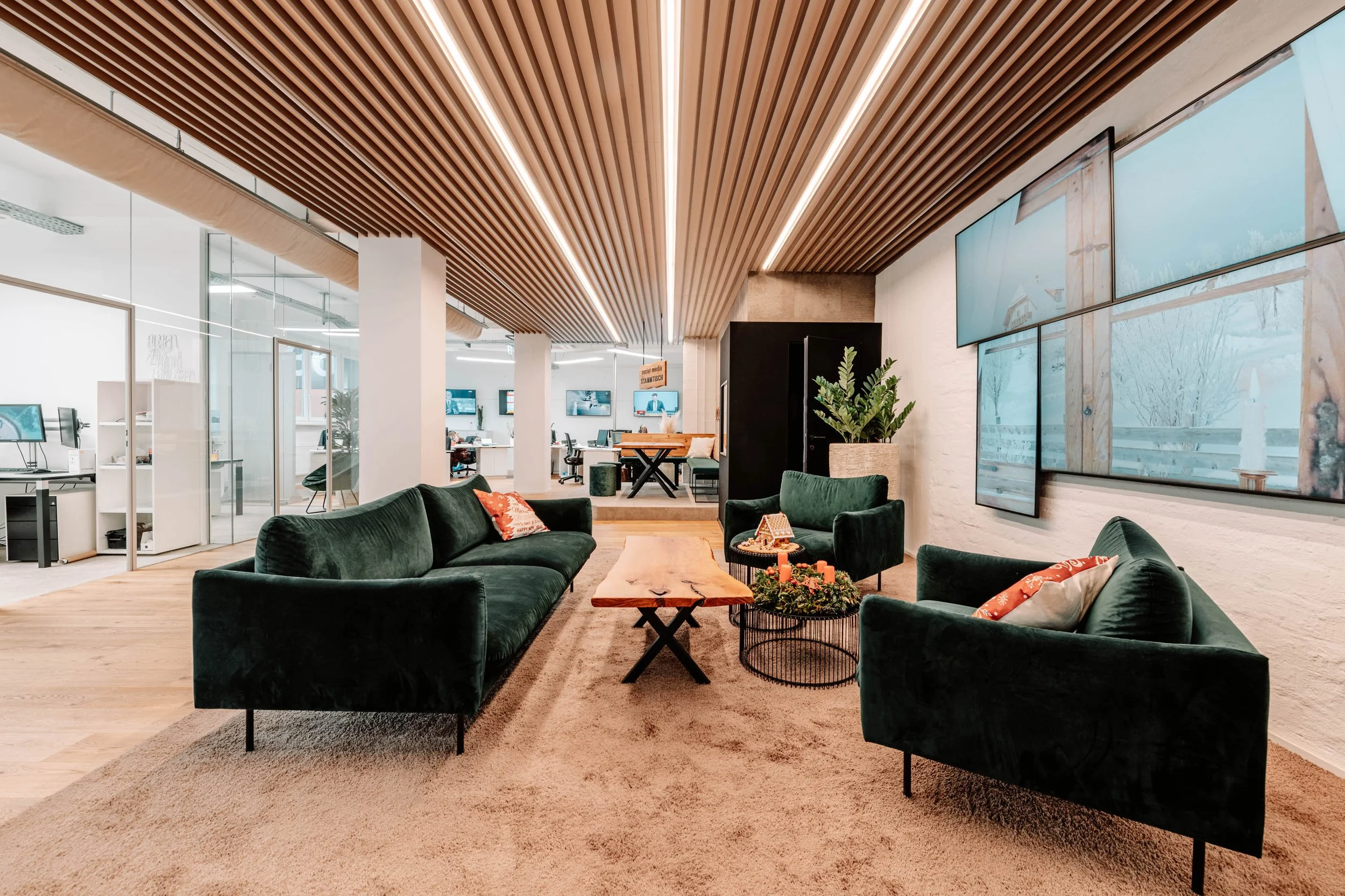 Moderne Lobby mit grünen Samtsofas, Holz-Tisch, großen Wandbildschirmen, Holzdecke und Pflanzen.