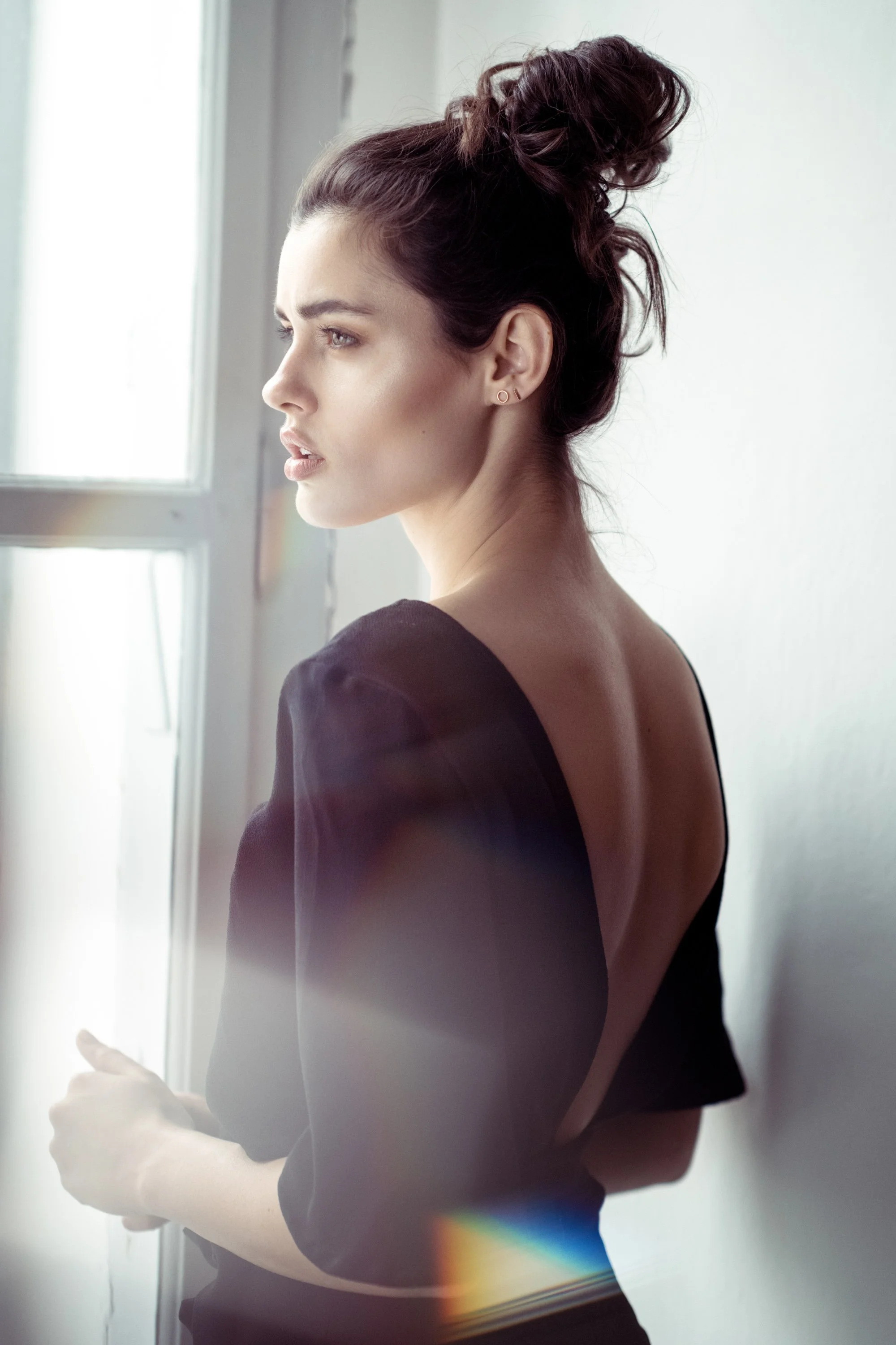 Eine junge Frau mit dunklem, hochgestecktem Haar schaut nach draußen, neben einem Fenster, tragt ein schwarzes Kleid mit offenem Rücken vor einer weißen Wand.