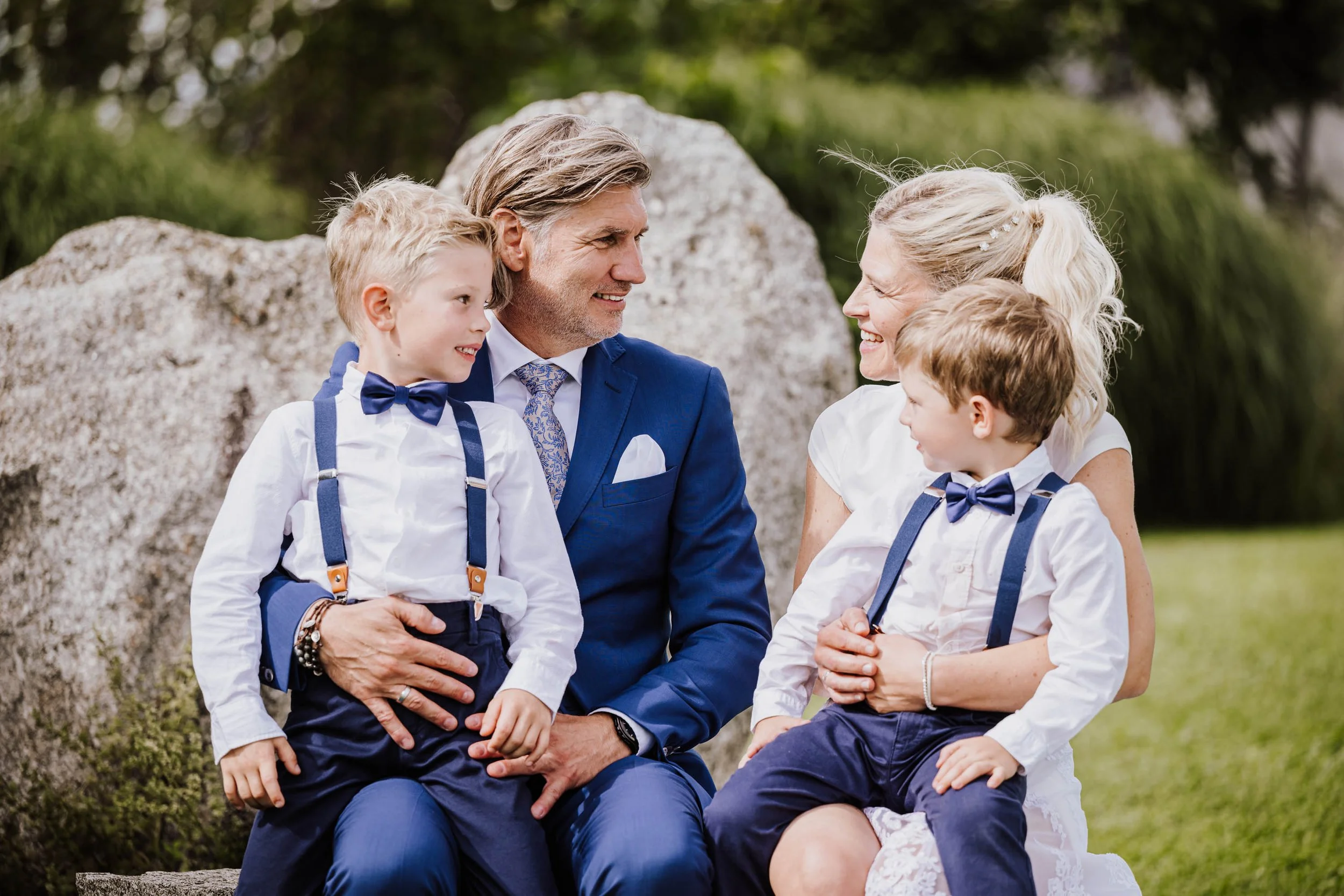 Eine glücklich aussehende Familie sitzt im Freien auf einem Stein, die Eltern lachen und schauen sich liebevoll an, während die beiden jungen Jungen mit weißen Hemden, Blauen Hosen und Fliegen auf den Elterngeschwistern sitzen, alle in formeller Klei