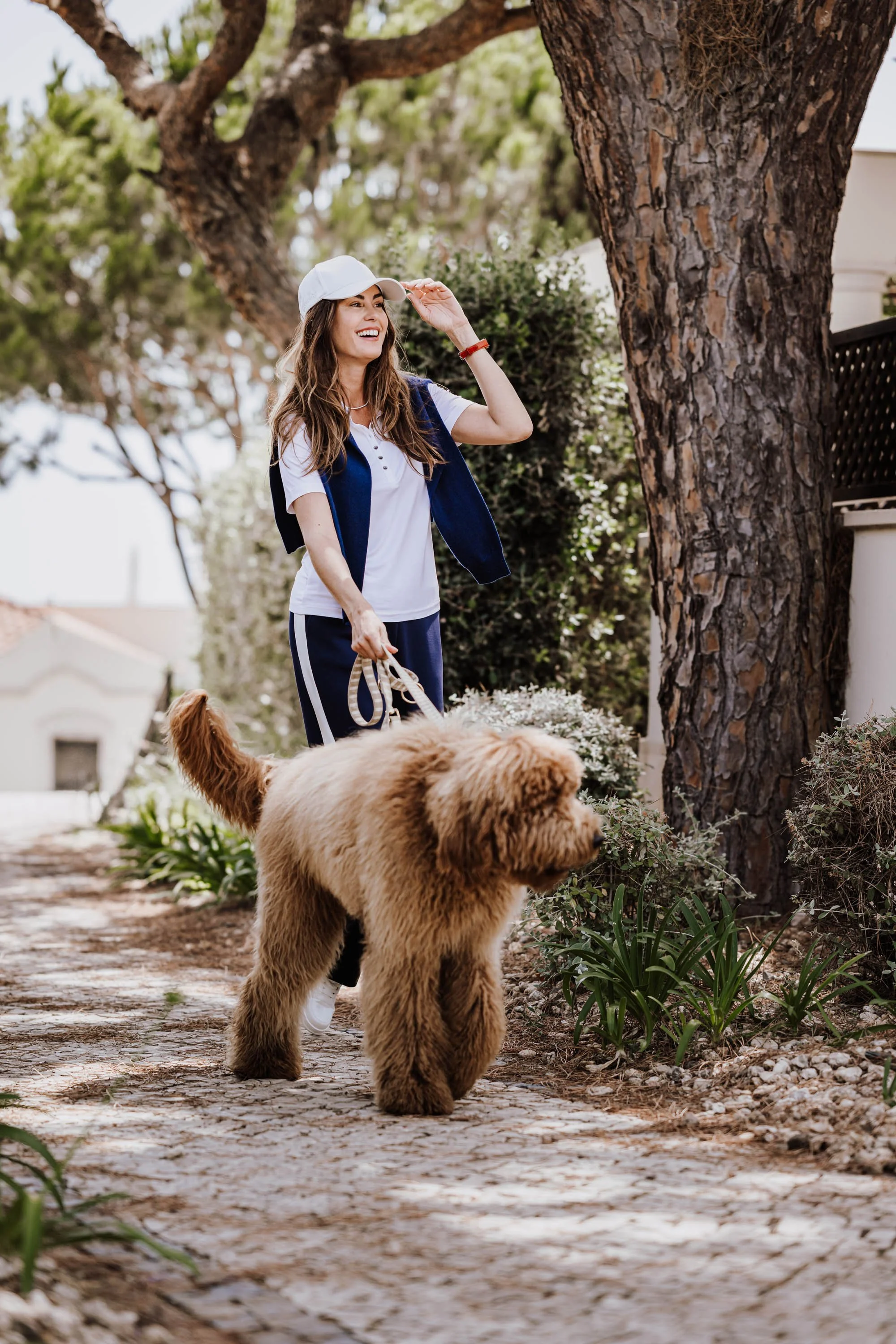 Eine lachende junge Frau mit einem Hund auf einem Weg im Freien, umgeben von Bäumen und Pflanzen.
