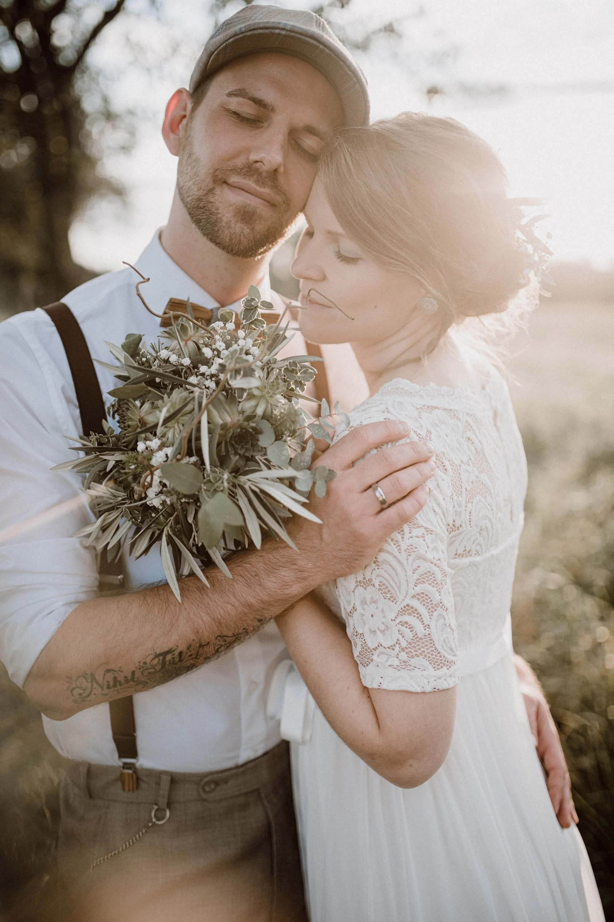 Ein Paar bei einer Hochzeitsaufnahme im Sonnenuntergang, die Frau trägt ein weißes Spitzenkleid, der Mann trägt Hemd mit Lederhosen, sie halten sich in einer ländlichen Umgebung.
