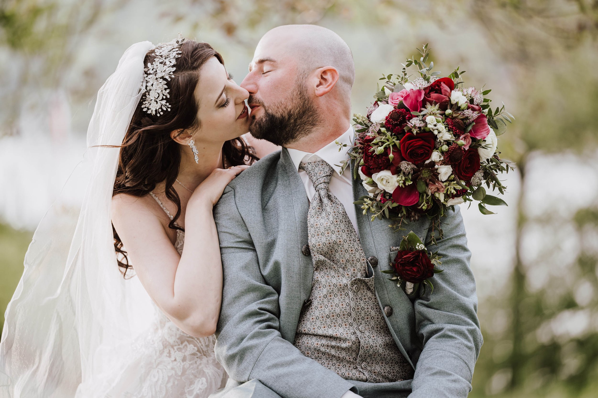 Ein Brautpaar, das sich bei einer Hochzeit küsst, mit der Braut in einem weißen Kleid und Schleier, und der Bräutigam in einem grauen Anzug mit Weste, im Hintergrund Bäume.