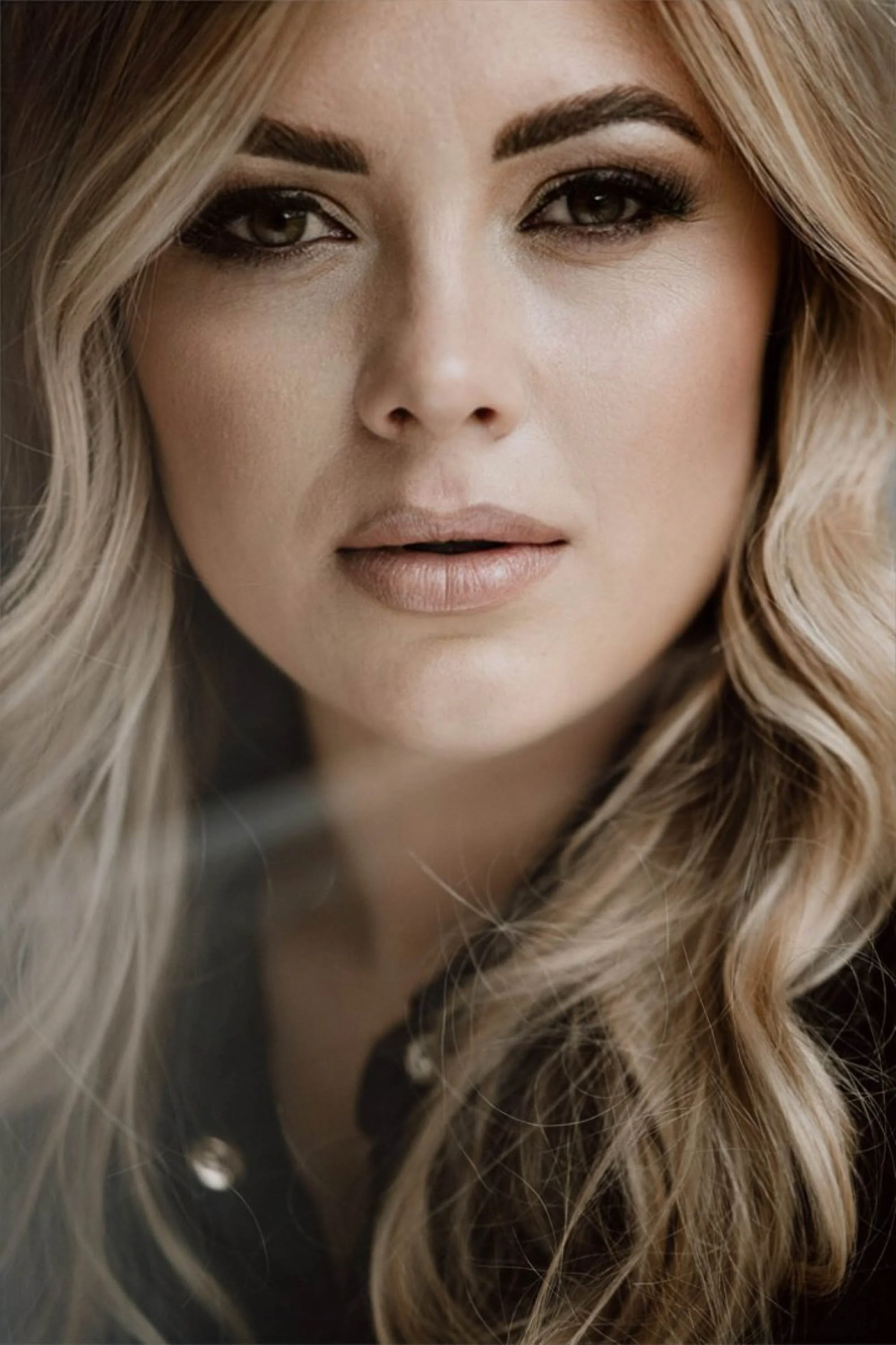 Nahaufnahme eines lächelnden Frauengesichts mit blonden, lockigen Haaren und makeup, in einem dunklen Oberteil.