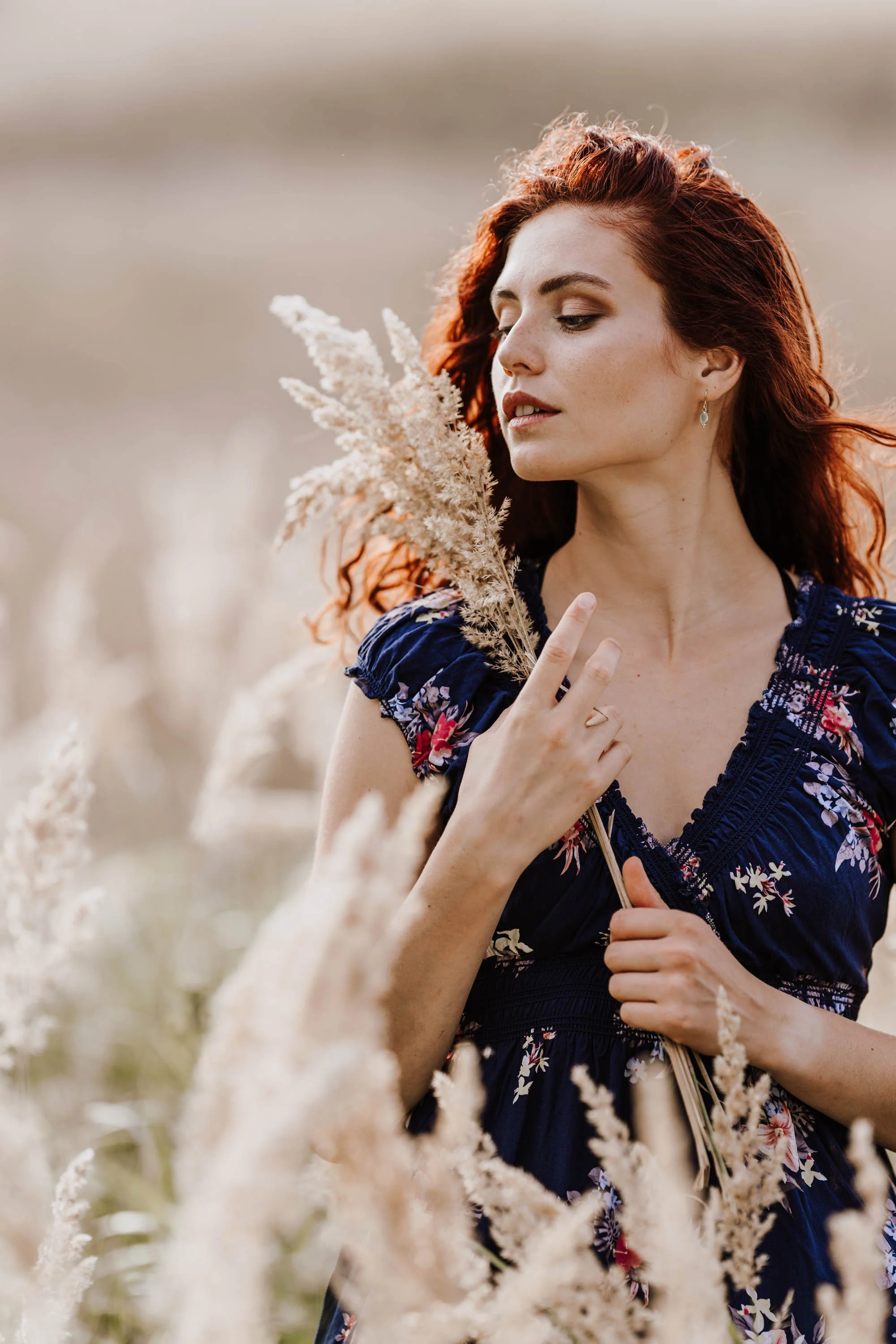 Eine junge Frau mit roten, lockigen Haaren in einem blauen, floral bedruckten Kleid steht in einem Feld mit getrockneten Pflanzen und hält eine Staude in der Hand.