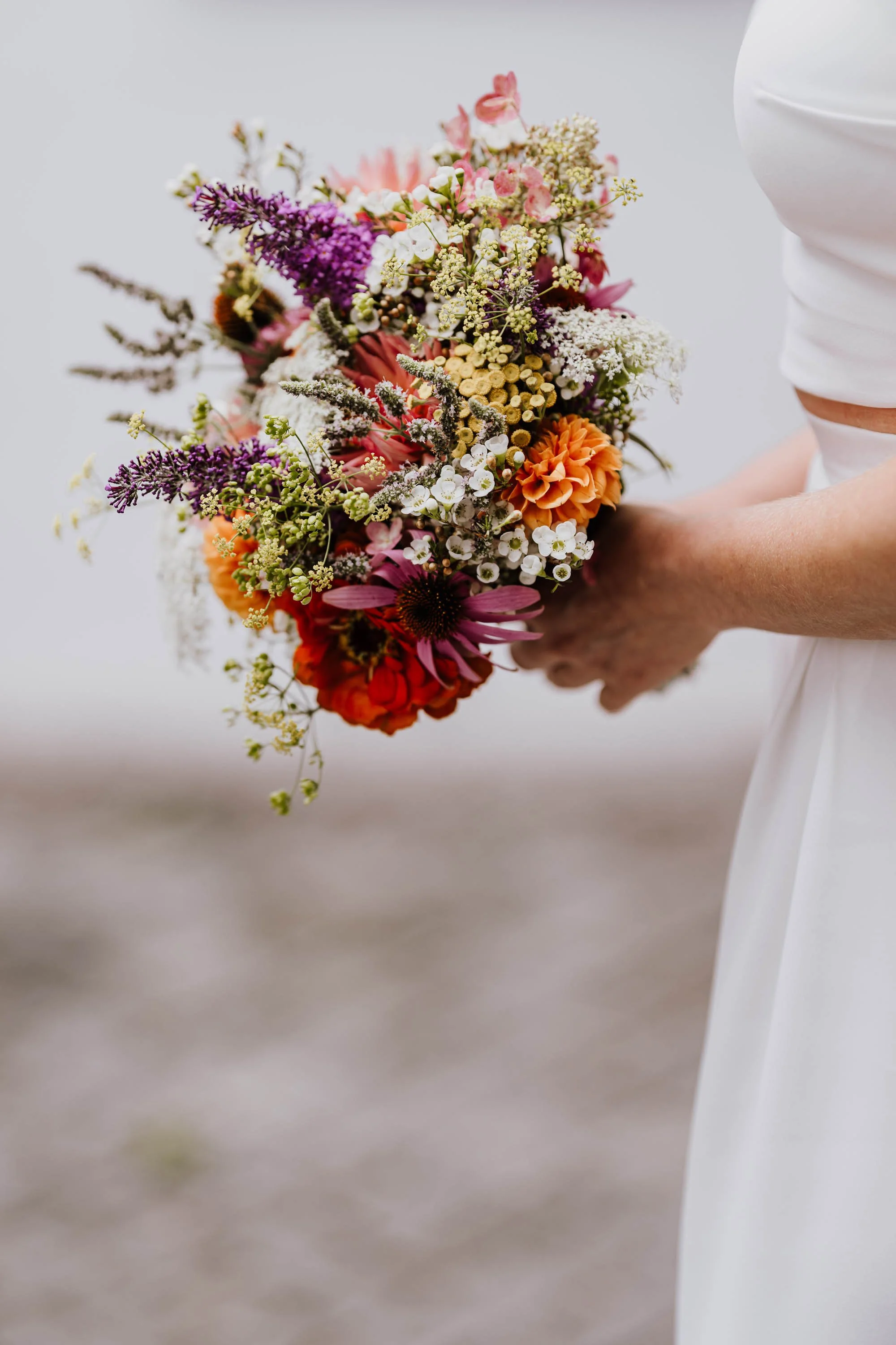 Hand hält einen bunten Hochzeitsblumenstrauß aus verschiedenen Blumen in Pastell- und Erdtönen, gehalten von einer Person in Weiß.