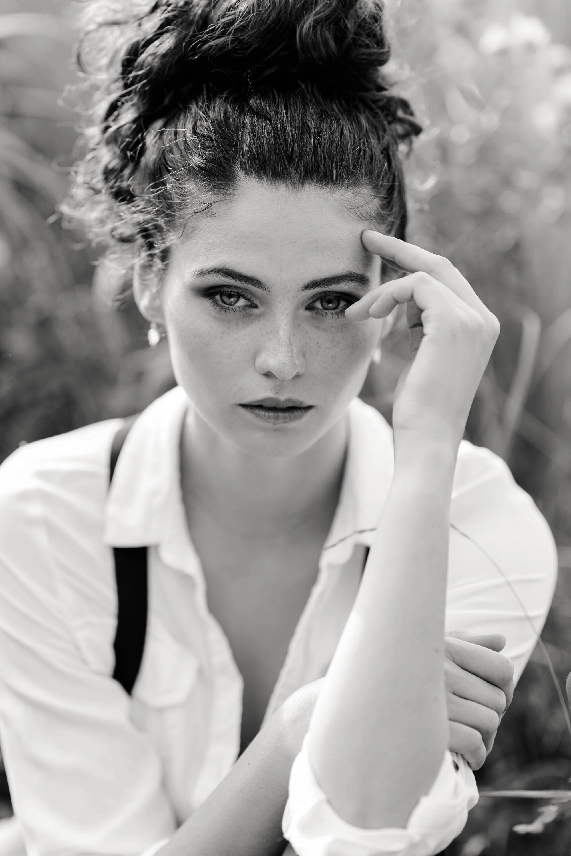 Schwarz-weiß-Fotografie eines jungen Mädchens mit hochgestecktem, lockigem Haar, das in die Kamera schaut, die Hand an die Stirn gelegt, im Freien mit verschwommenem grünem Hintergrund.