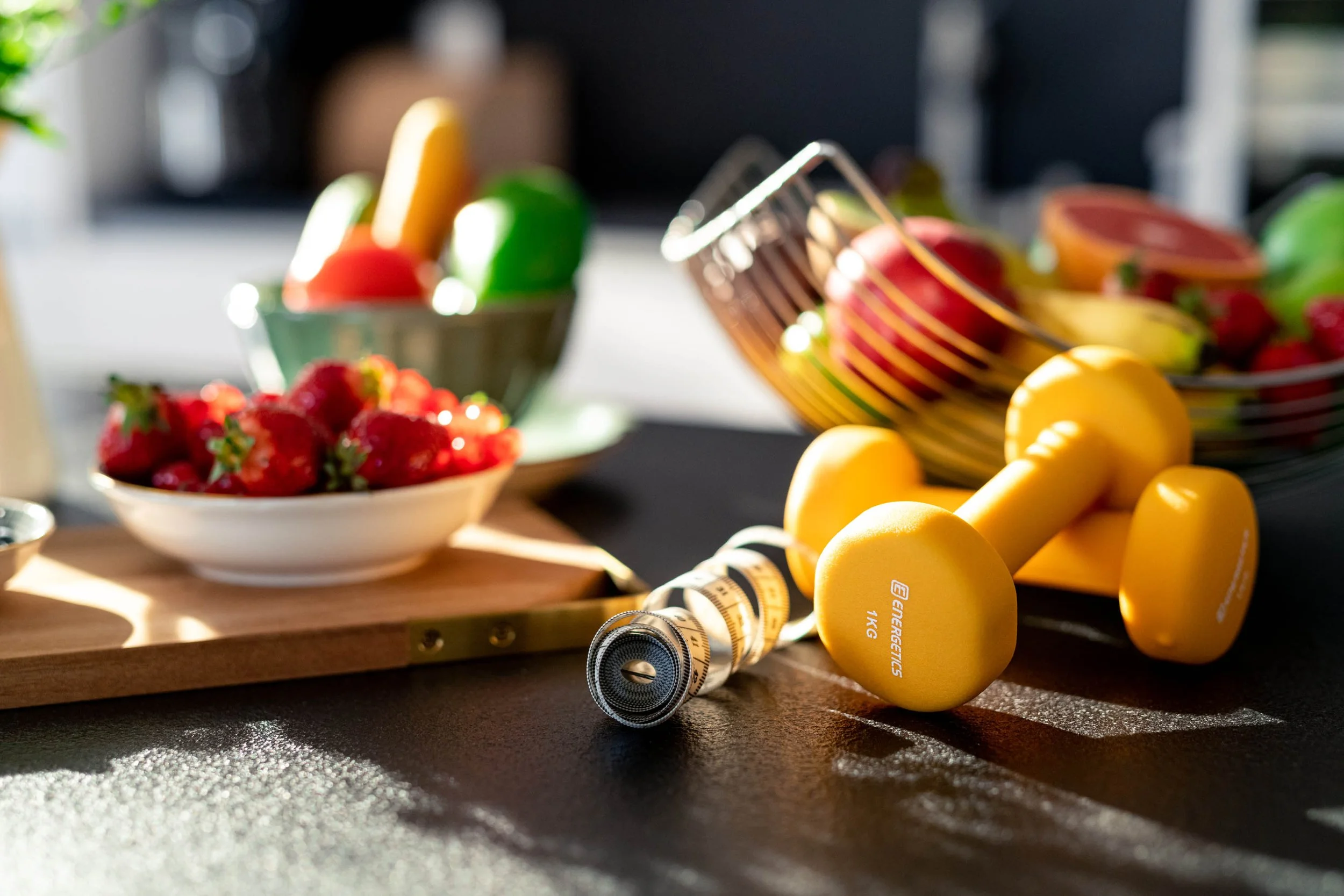 Gelbe Hanteln, ein Maßband, frische Erdbeeren, bunte Äpfel und weitere Früchte auf einer schwarzen Oberfläche bei Sonnenlicht.