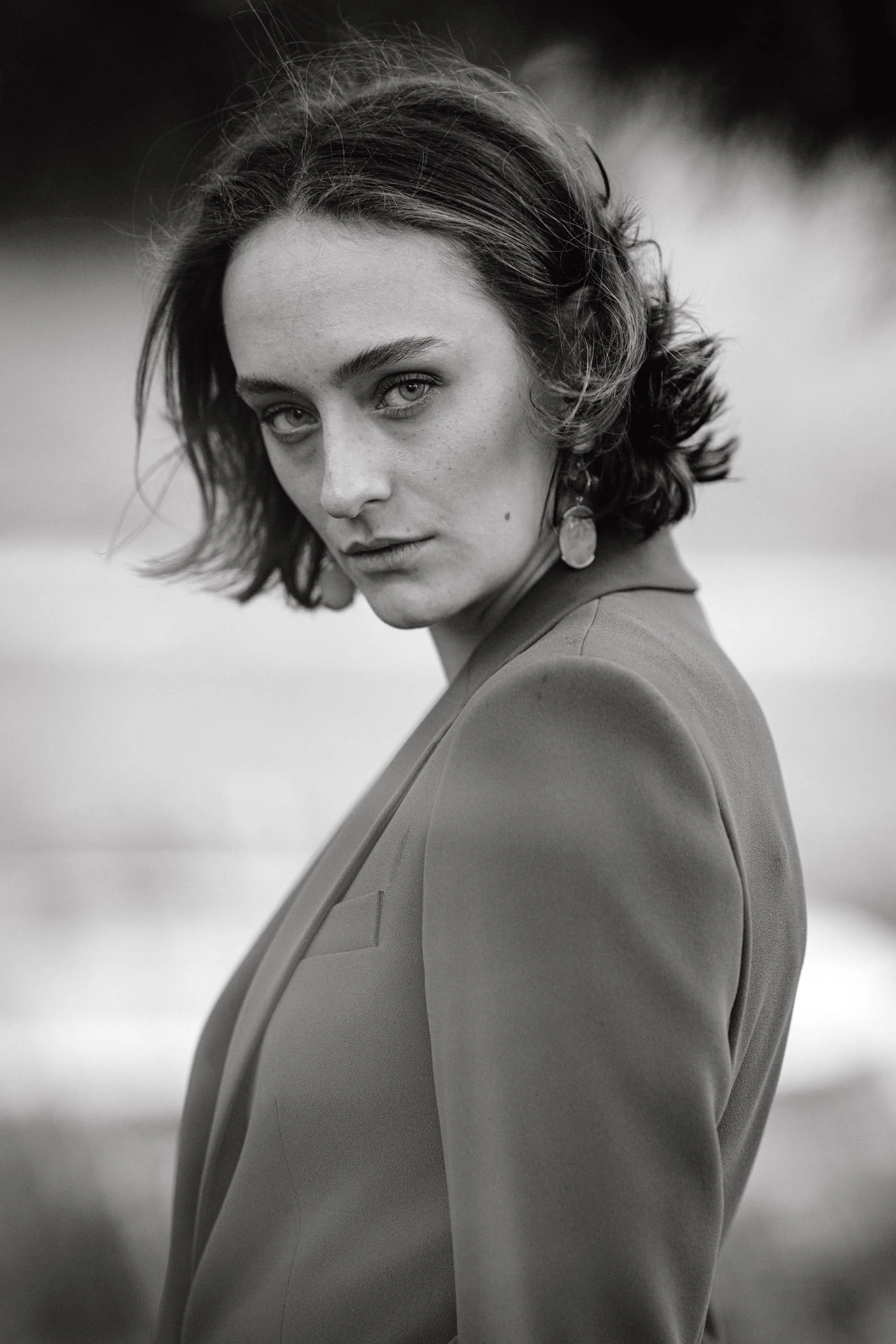 Schwarz-Weiß-Foto einer jungen Frau mit kurzen, gewellten Haaren, die eine ernste Miene zeigt, vor unscharfem Hintergrund, trägt einen Blazer und große Ohrringe.