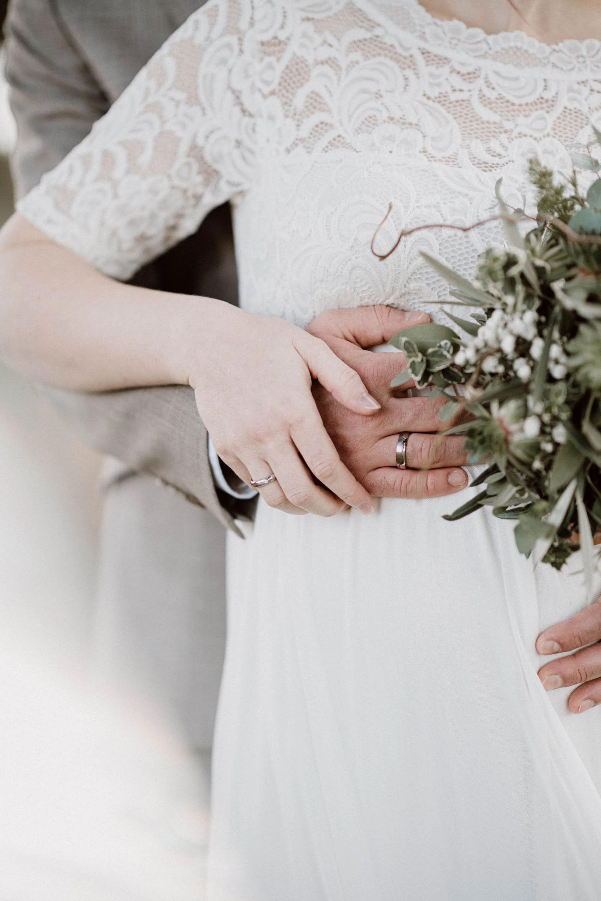 Nahaufnahme eines Brautpaares, das sich an der Taille umarmt, mit focus auf die Hände und Eheringe. Die Braut trägt ein weißes Kleid mit Spitzenärmel, während der Bräutigam einen grauen Anzug trägt.